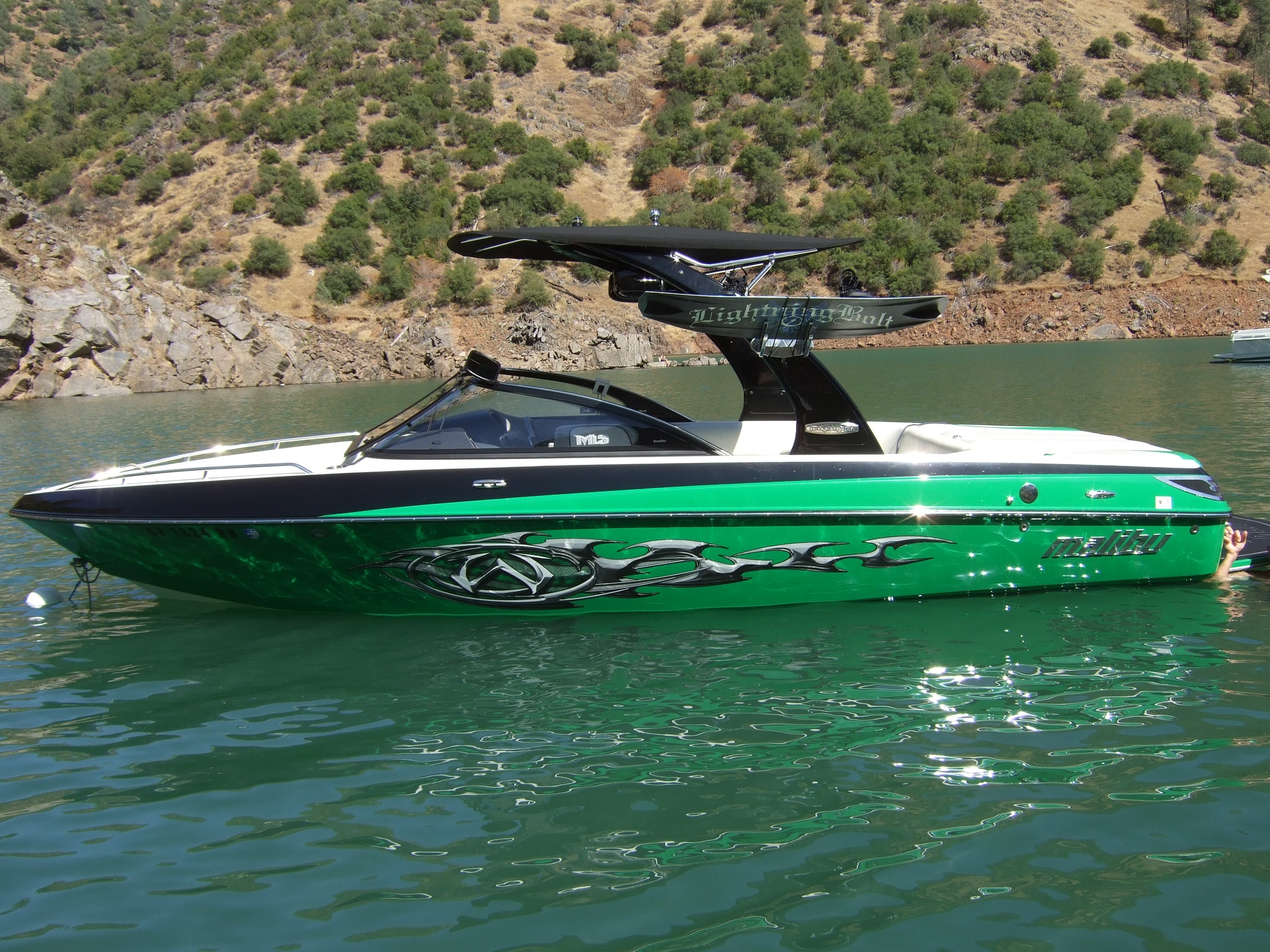 Malibu Illusion X Bimini top installed on a green and black Malibu boat on calm water with rocky hills in the background.