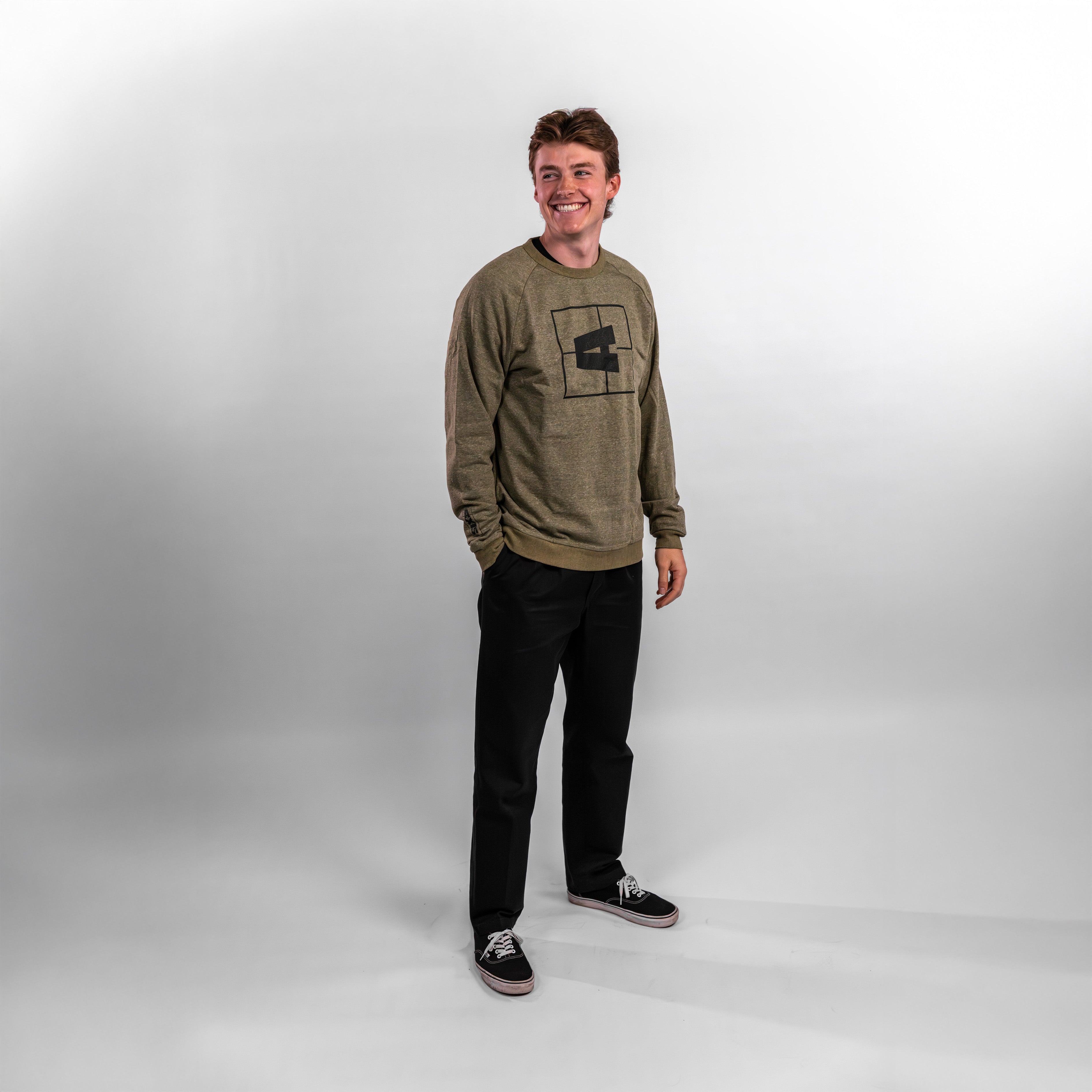 A young man stands smiling in a casual olive green Axis crewneck sweatshirt paired with black pants and sneakers.