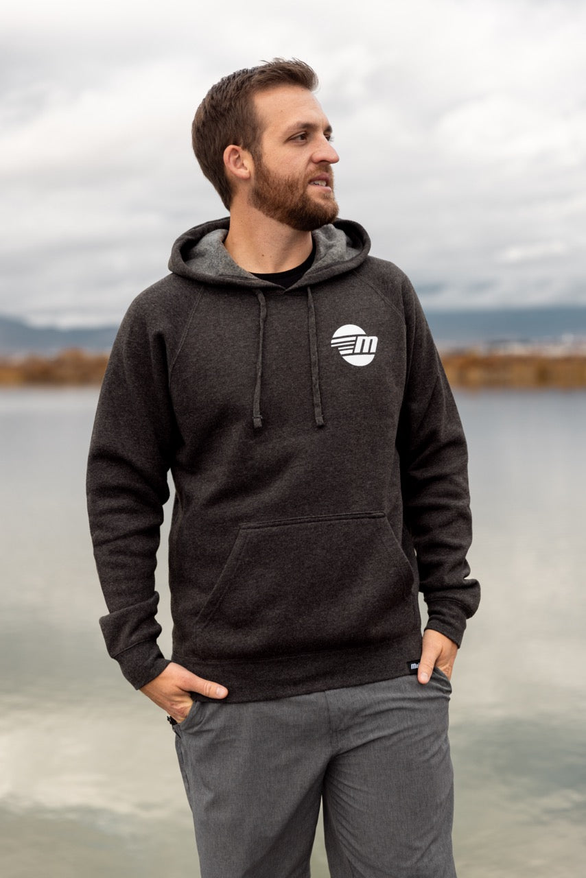 Man outdoors wearing a dark gray Malibu Hoodie with a white circular logo, hands in pockets, near a body of water.