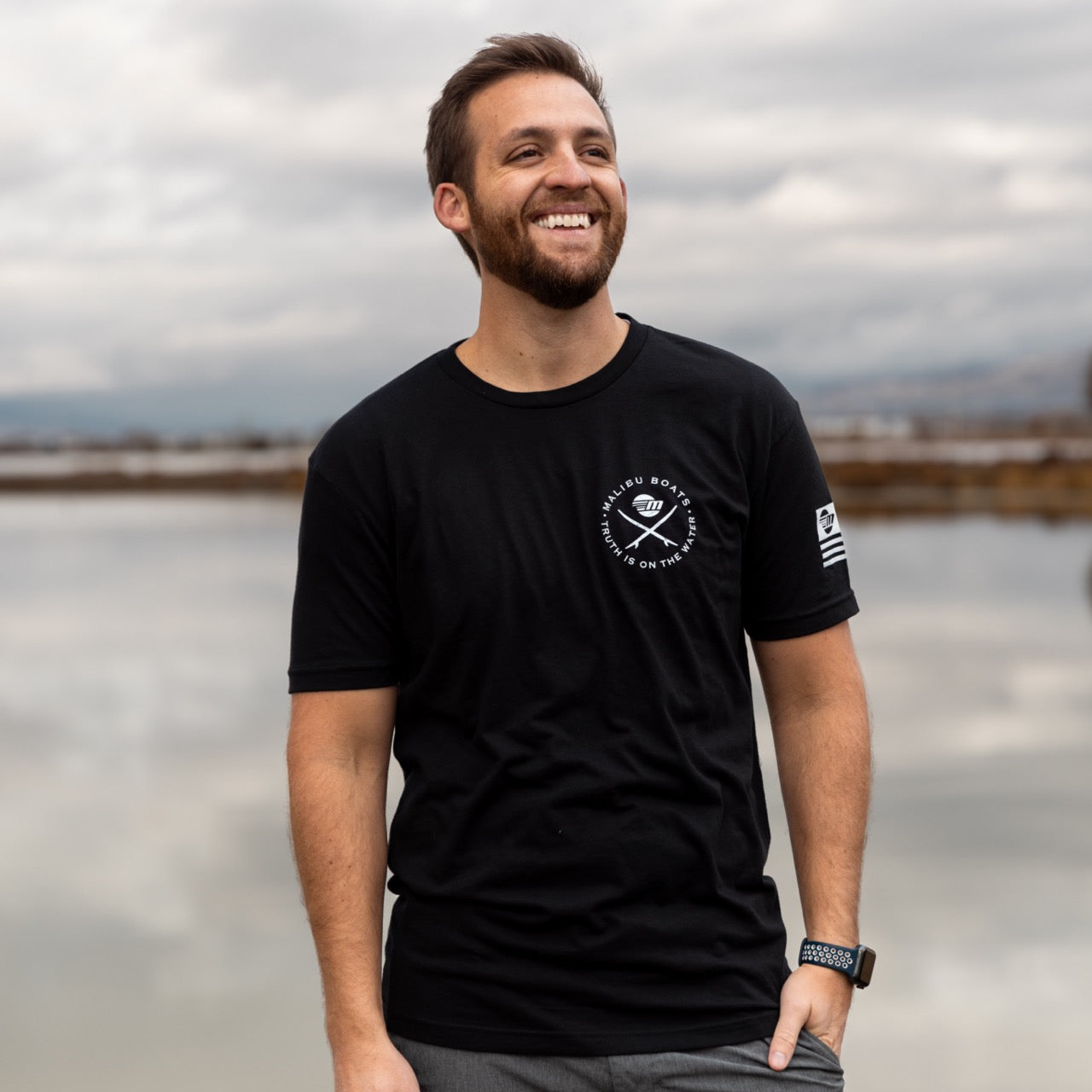 Man smiling outdoors wearing a black Malibu Endless Wake Tee with white logo and text, standing near water on a cloudy day.