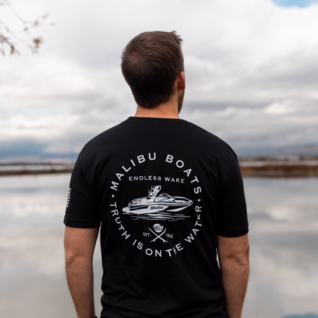 Back view of a man wearing a black Malibu Endless Wake T-shirt with boat graphic and water-themed text, standing outdoors near water.
