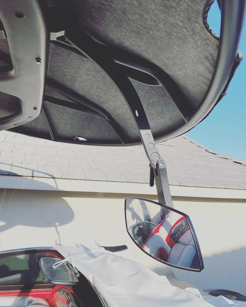 Underneath view of the Mirror Tower Arm-Illusion X, a black adjustable mirror arm attached to a vehicle's interior, with a car's side mirror and roof visible.