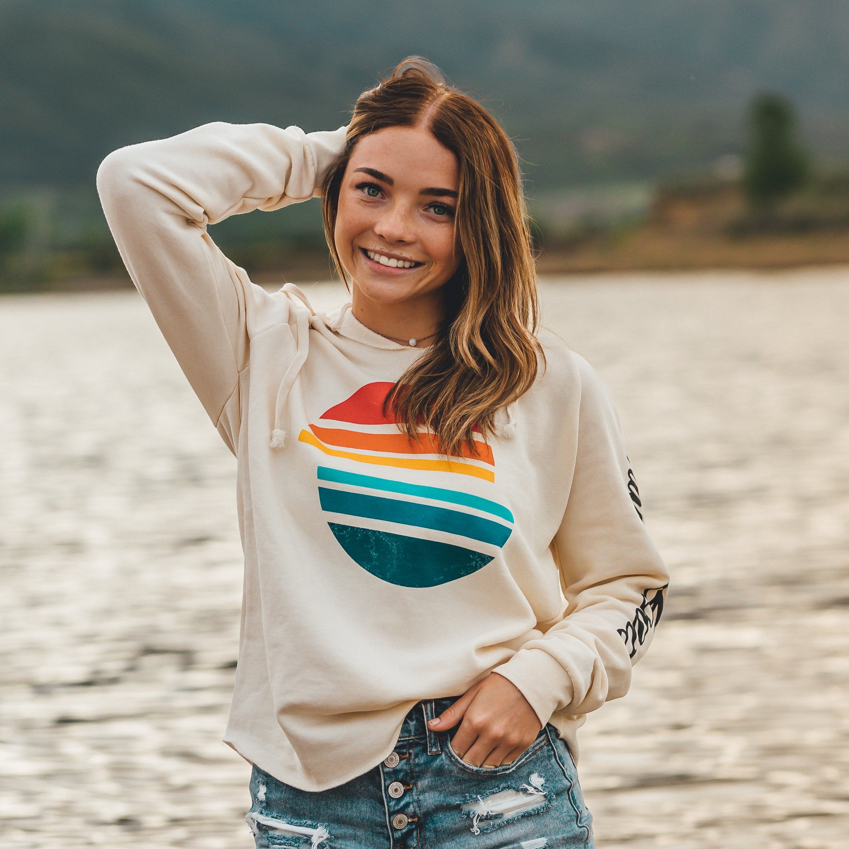 Woman smiling outdoors in a Sunsetter Pullover, featuring a colorful sunset graphic, near a lake with mountains in the background.