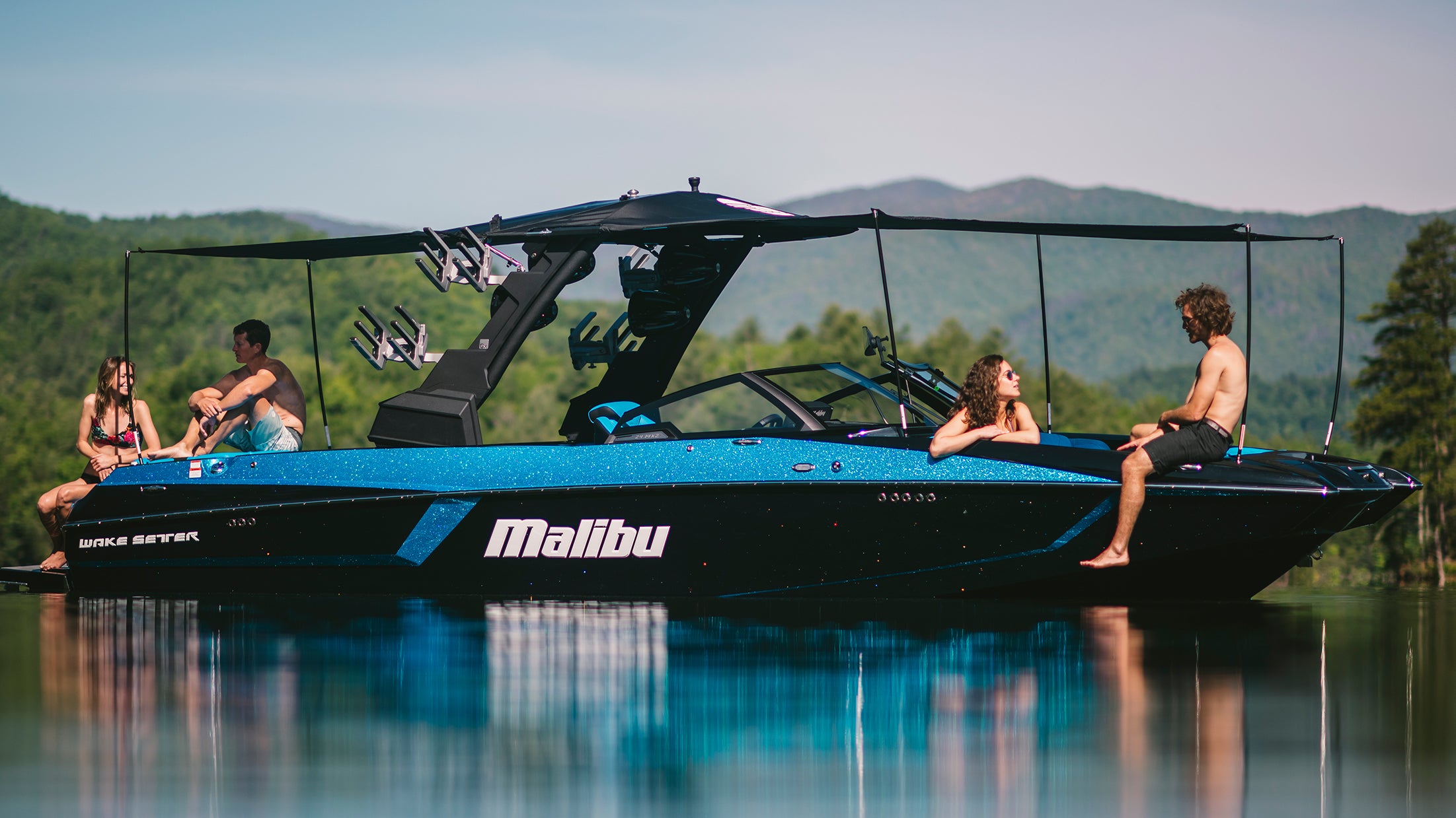 A blue Malibu 24MXZ Solo Mesh Awning boat with a G3 Tower, 20' length, on calm water with people relaxing onboard.
