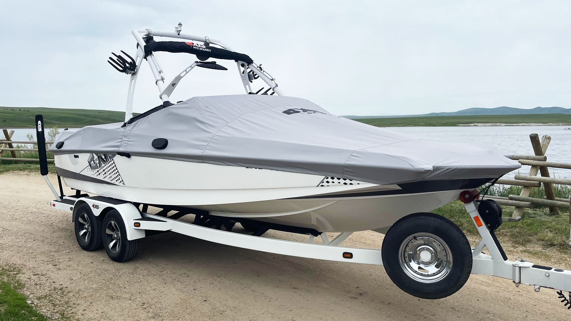 A white boat on a trailer with a gray cover, parked outdoors near a lake, featuring a tower labeled "FatAX Tower" for water sports.