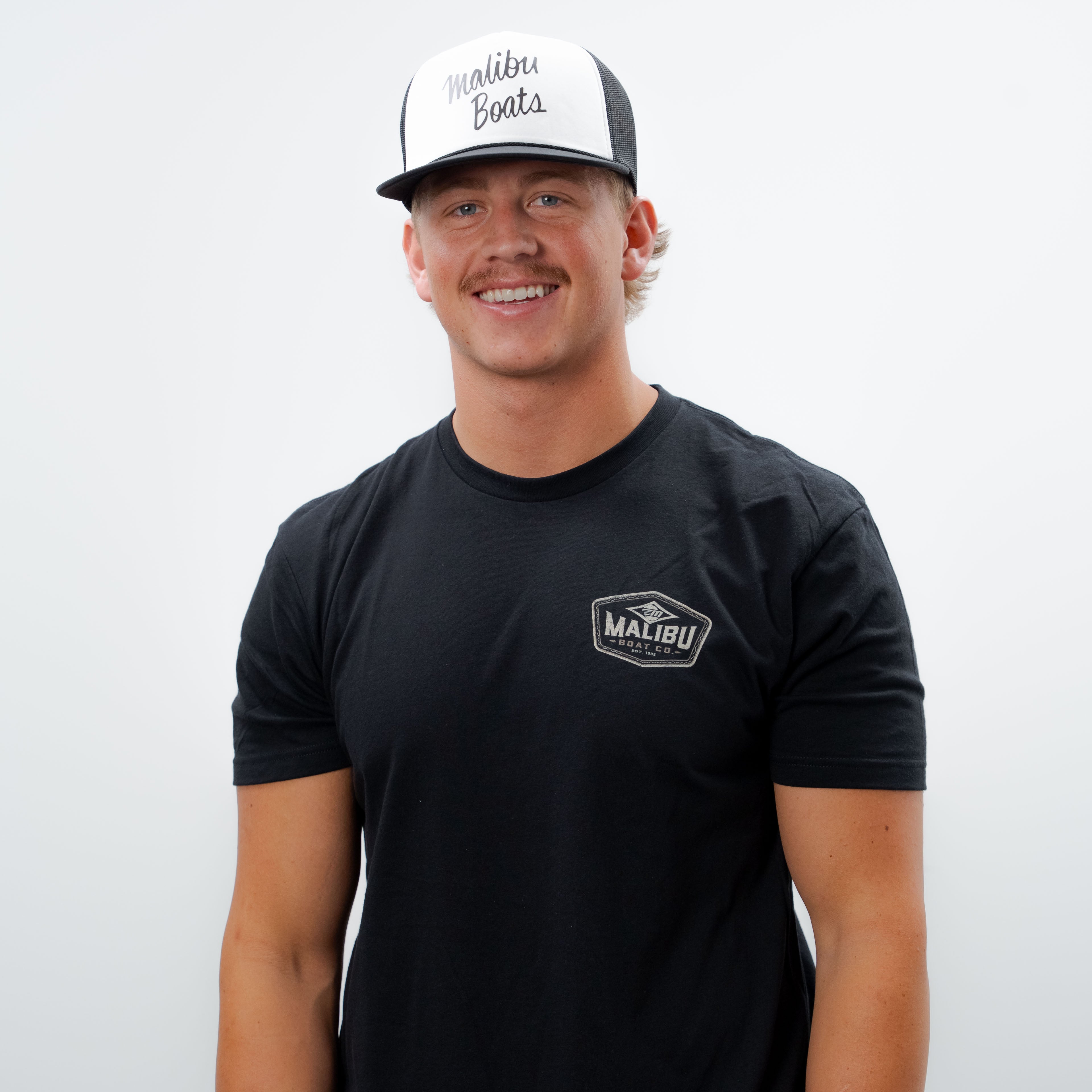 Man wearing a black Malibu Factory Trucker Hat with mesh back and front logo, smiling against a plain white background.