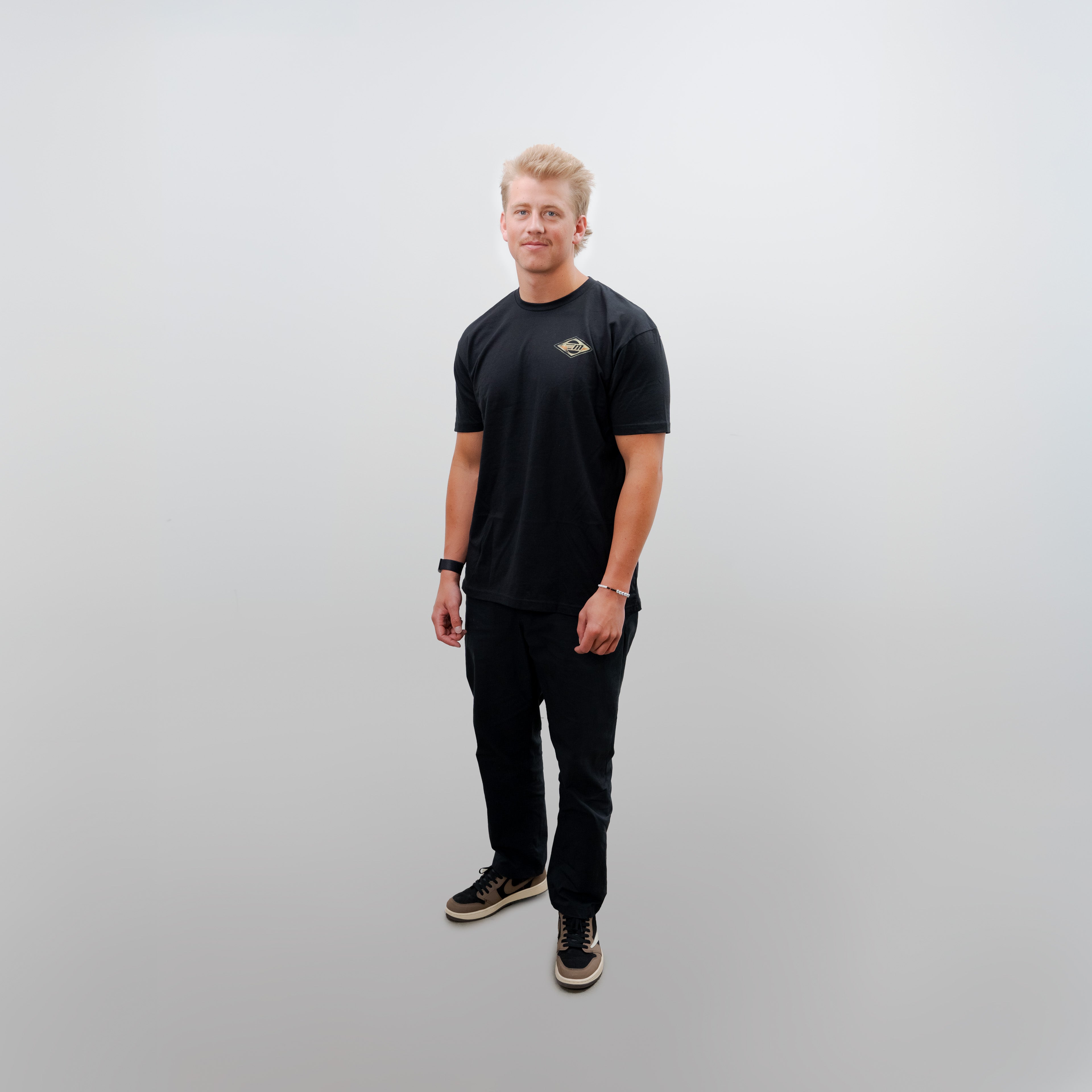 Young man wearing a black Malibu Surfer Tee, black pants, and sneakers, standing against a plain white background.