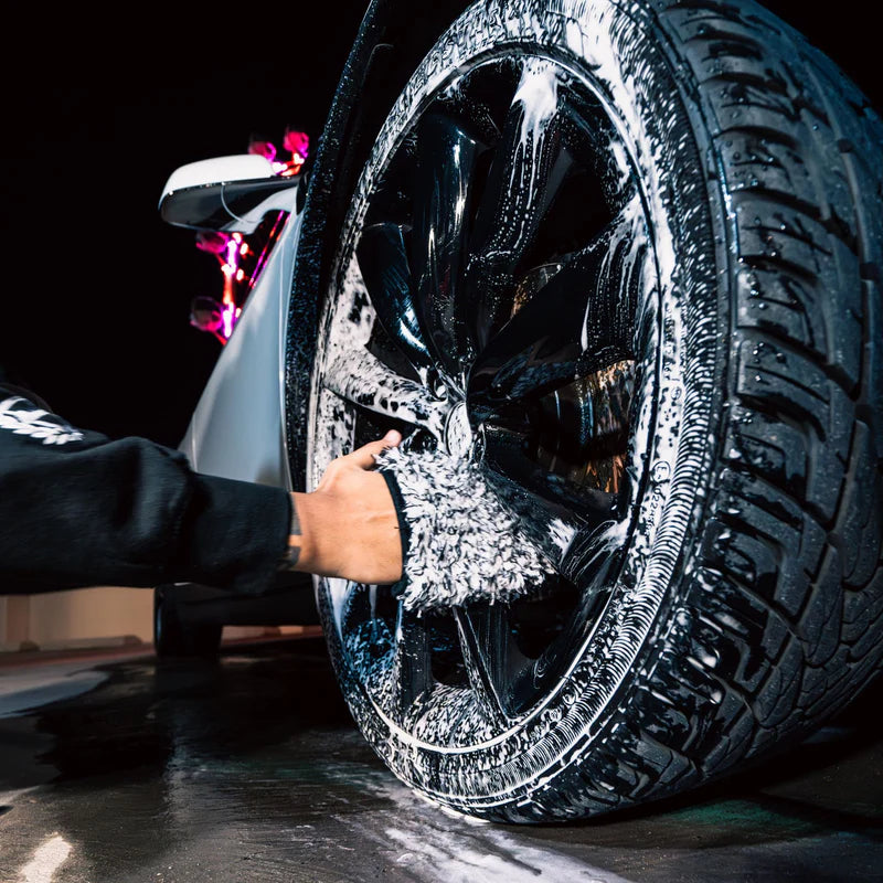 A person washing a car wheel with a microfiber wash mitt, creating foam and cleaning the black alloy rim effectively.