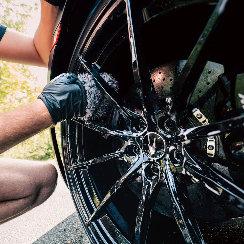 Person cleaning a shiny black car wheel with a microfiber wash mitt, wearing a black glove for effective dirt removal.