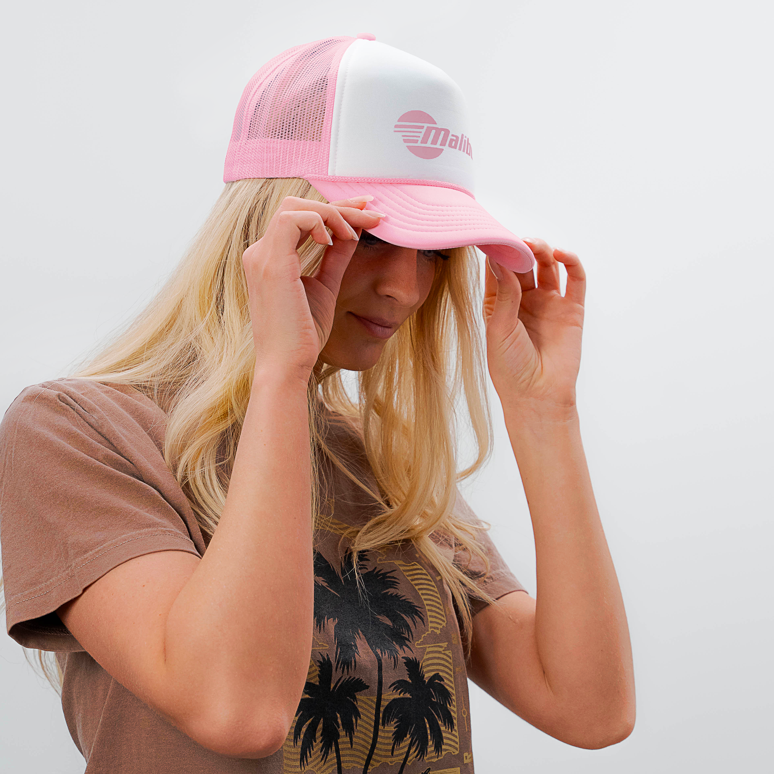 Woman adjusting a pink and white trucker hat with a "Malibu" logo, casual summer style, against a plain white background.