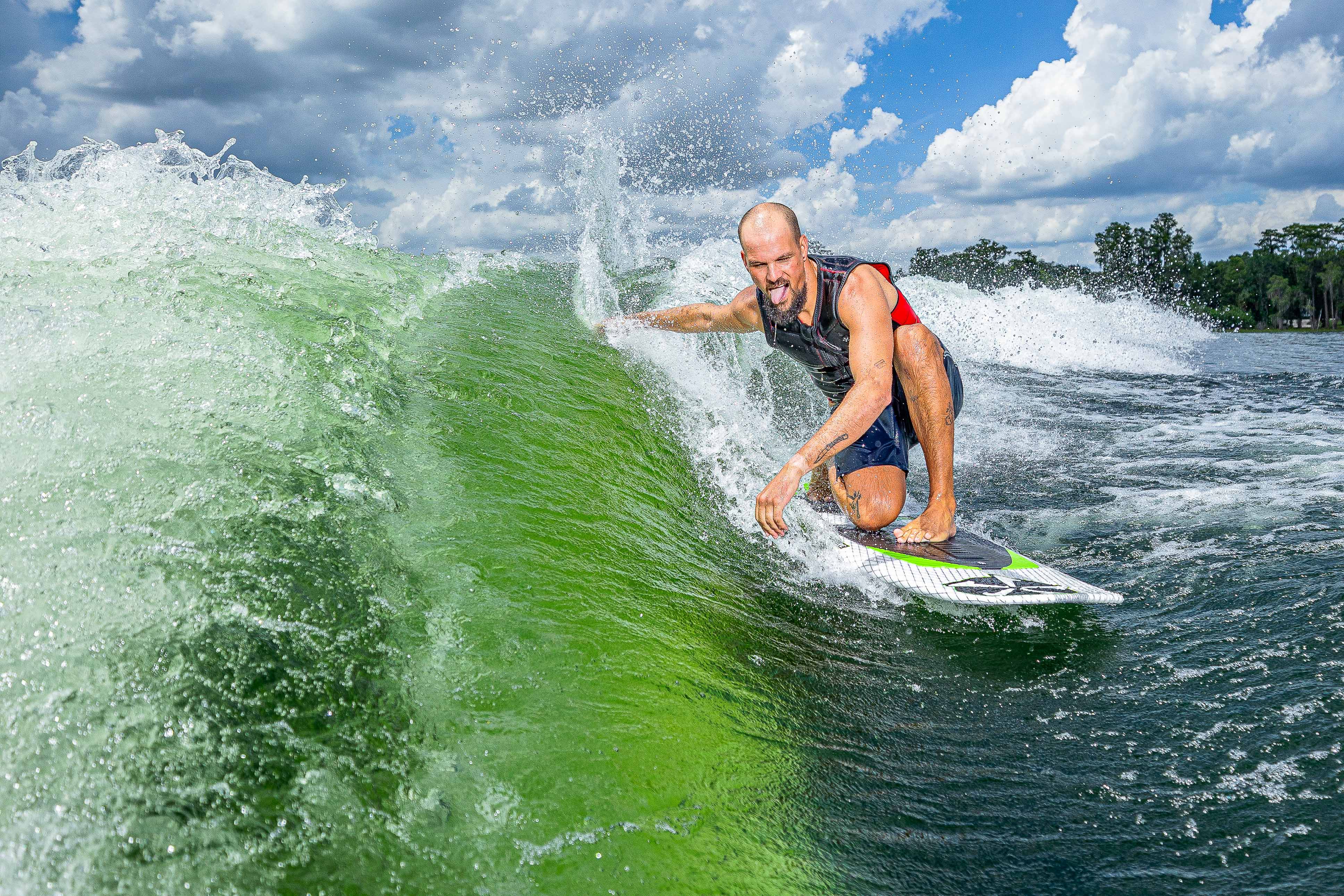 A man surfing on a green wave with a clear sky and trees in the background, riding a 2025 Phase 5 Doctor surfboard.