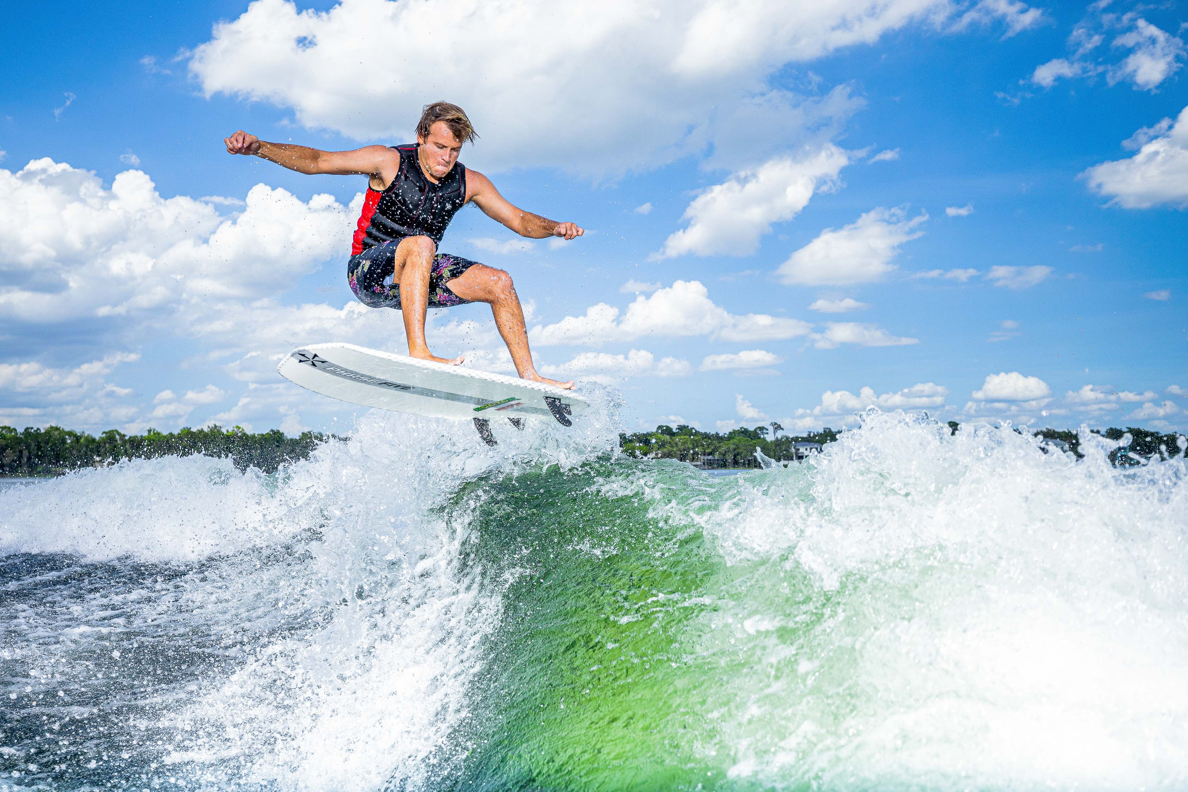 A person surfing on a white surfboard in the ocean, jumping over a wave under a blue sky with scattered clouds.
