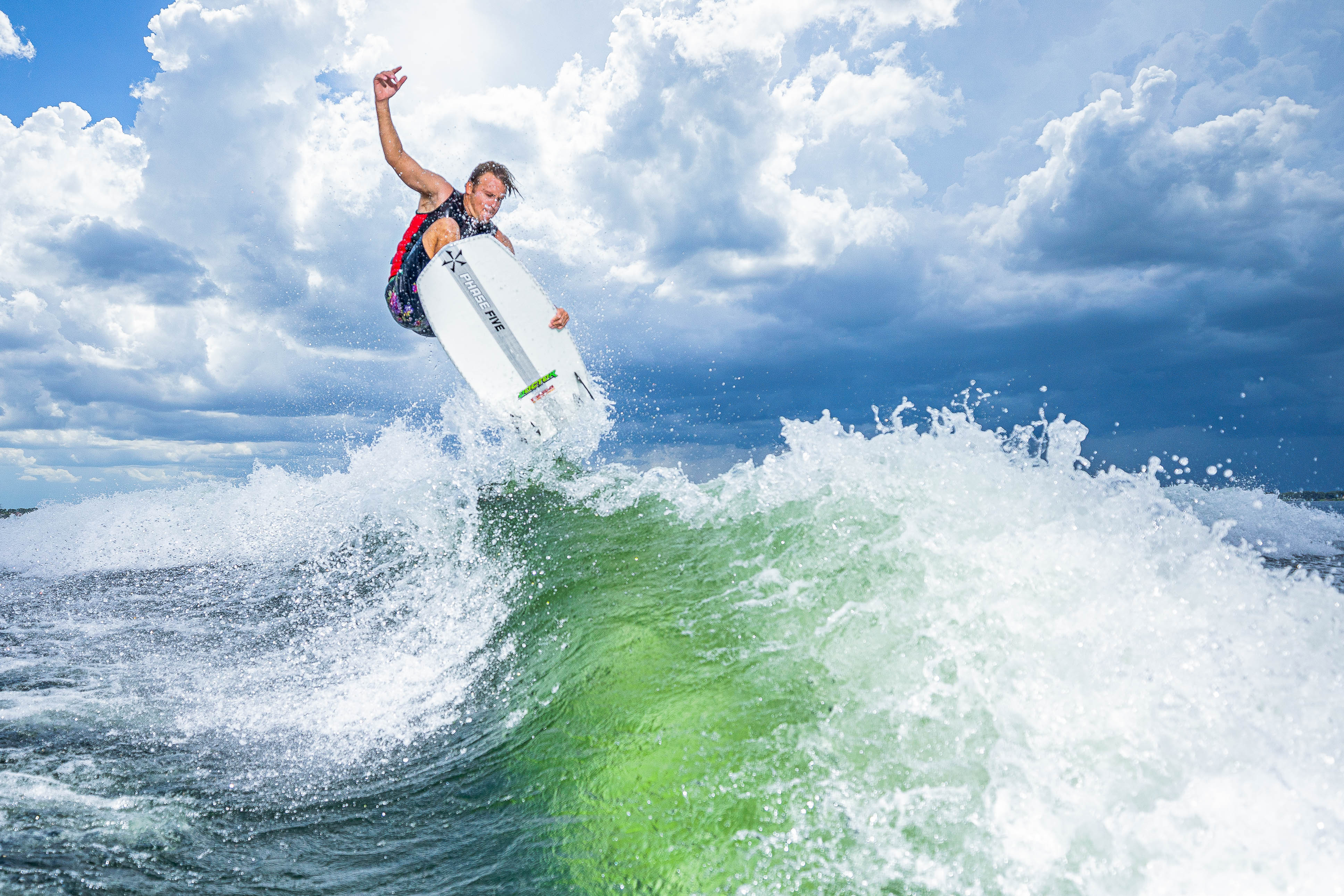 A surfer performs an aerial trick on a white Doctor Surf Board amid green waves and a cloudy sky.