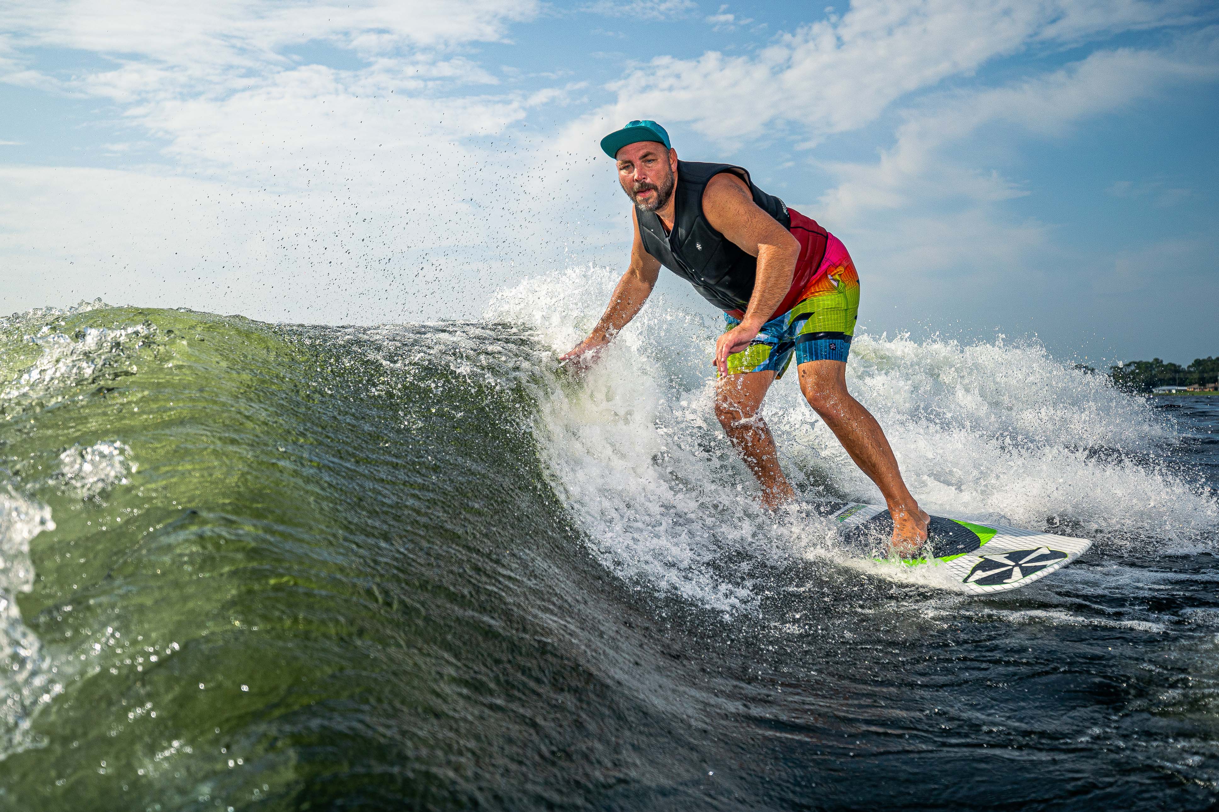 A man surfing on the 2025 Phase 5 Doctor Surf Board, riding a wave with a blue sky background and shoreline in distance.