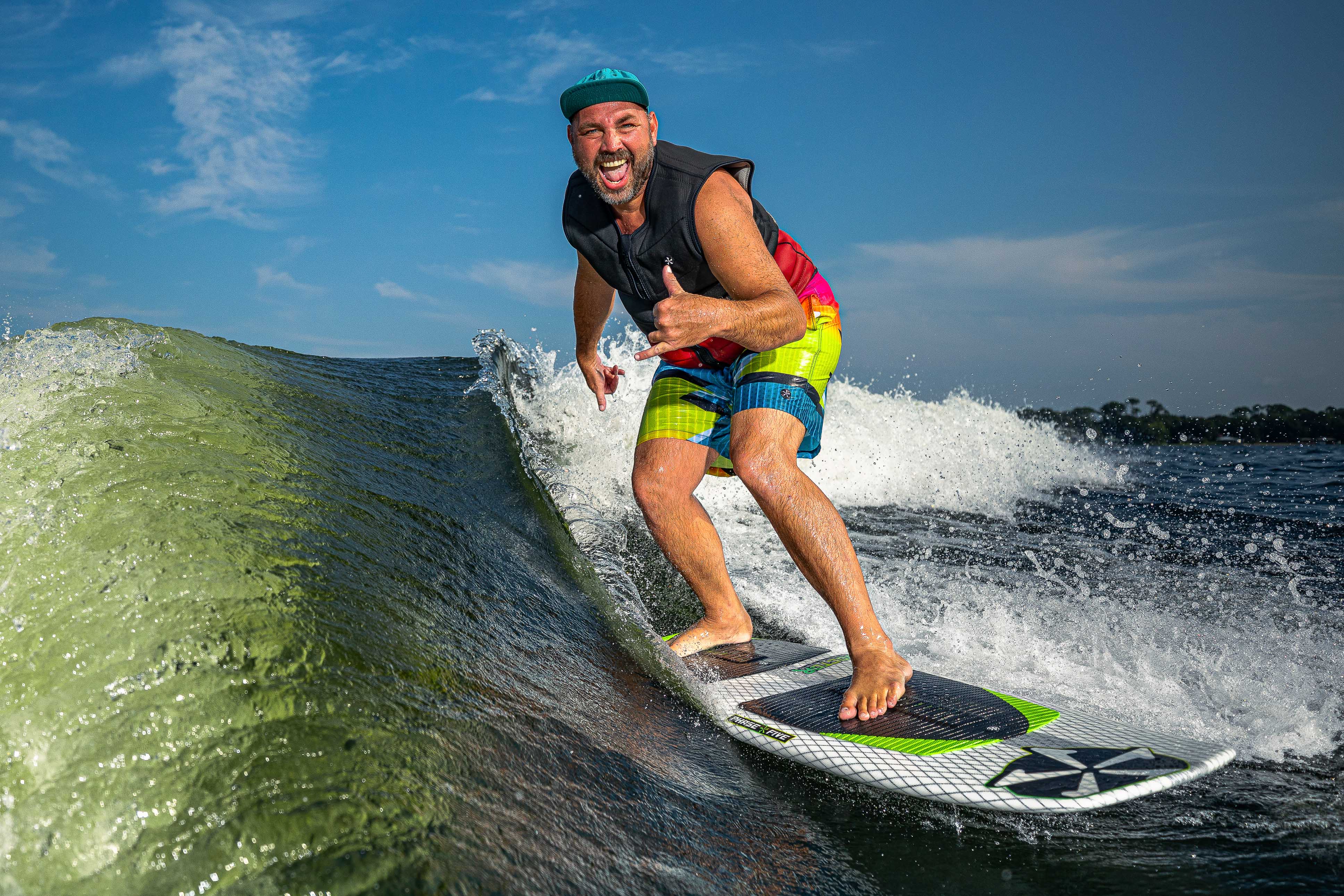 A man joyfully rides a wave on a 2025 Phase 5 Doctor Surf Board, showcasing vibrant swim trunks and a life vest.