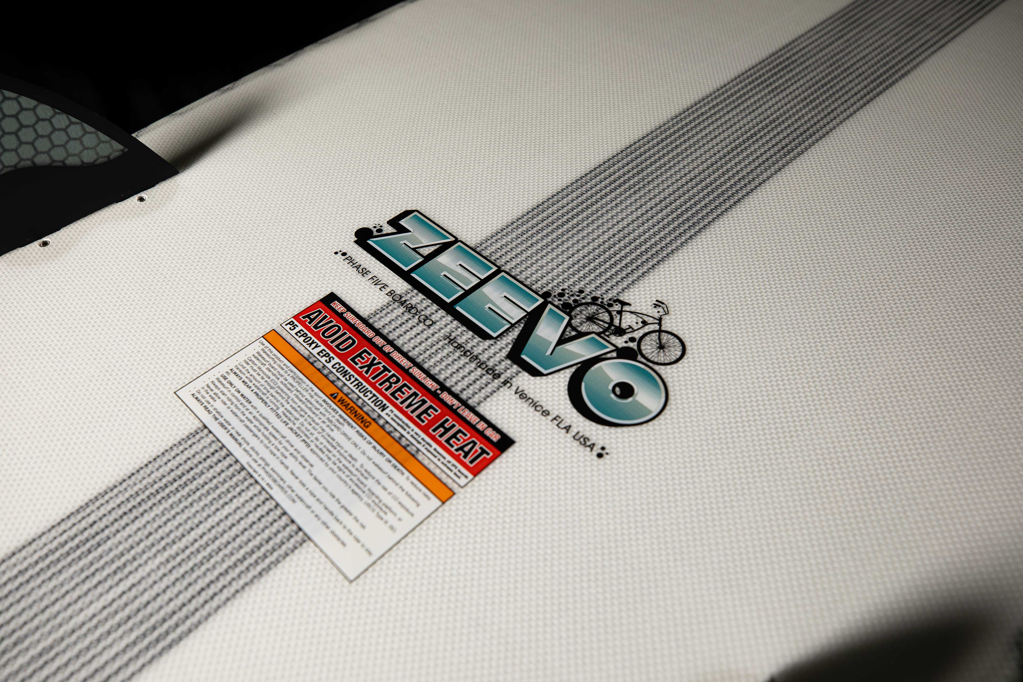 Close-up of the 2025 Phase 5 Zeevo surfboard with branding, a bicycle graphic, and warning labels on a textured white surface.