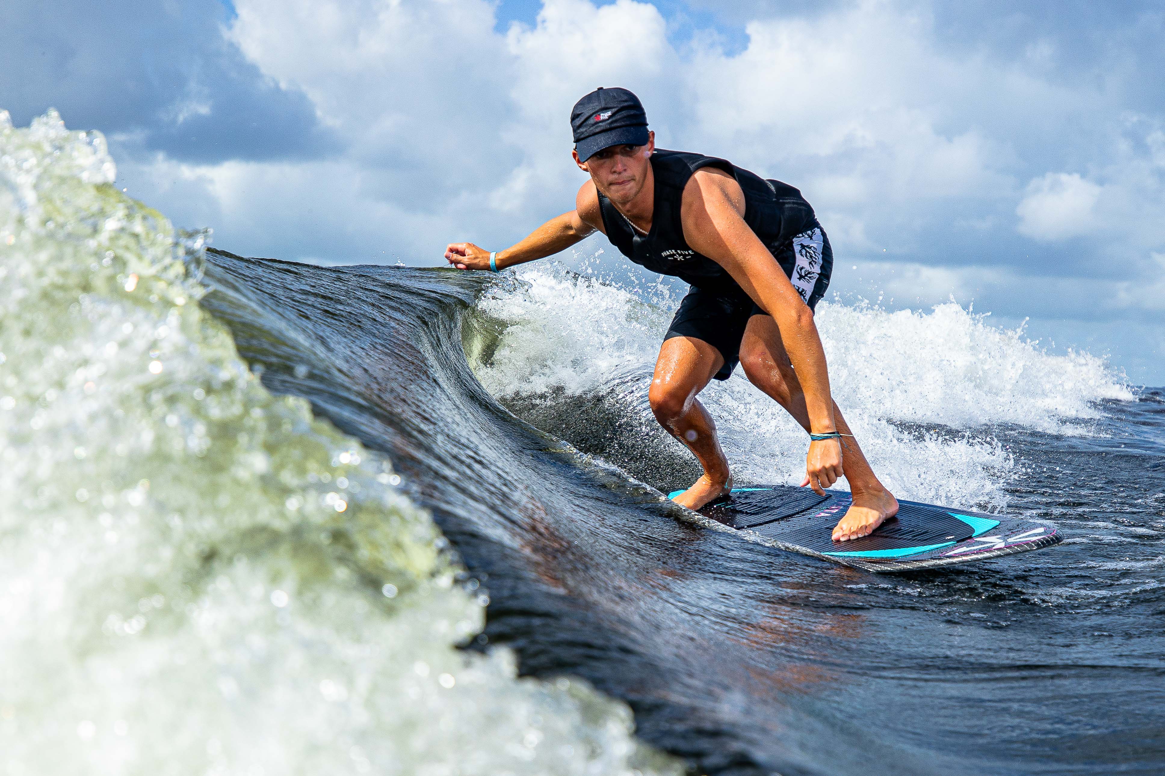 A person rides a wave on a 2025 Phase 5 Hypsta skim board, showcasing skill and balance in a sunny outdoor setting.