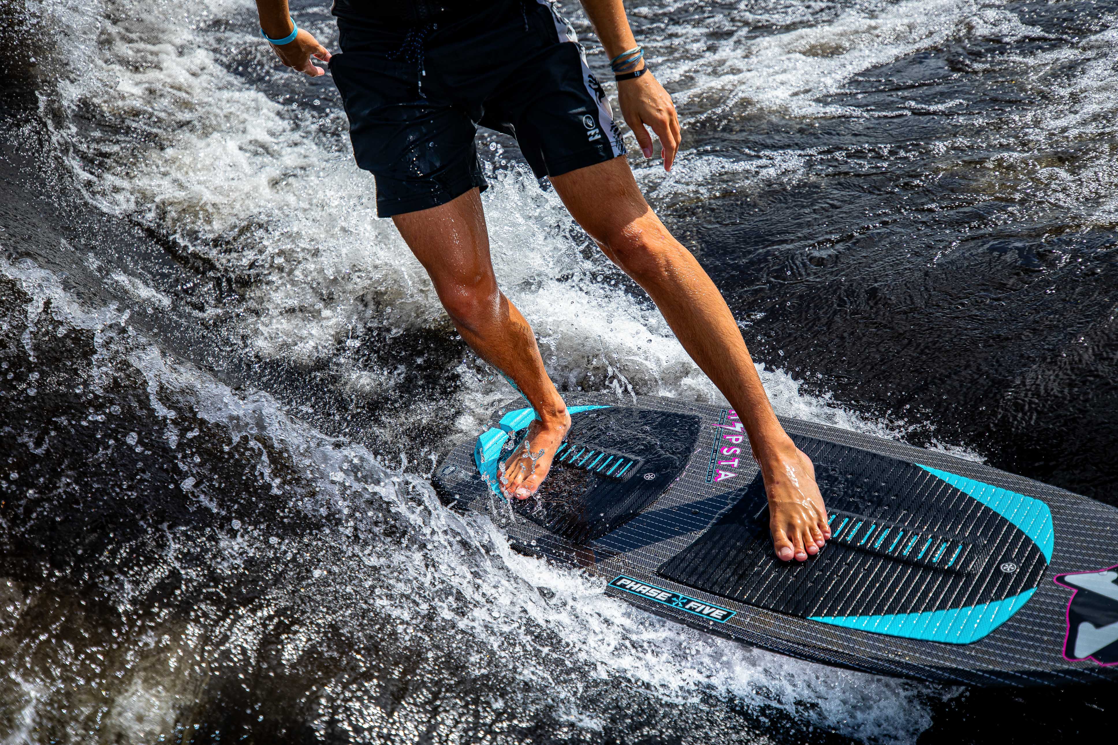 A person rides the 2025 Phase 5 Hypsta skim board on water, showcasing dynamic movement and splashing waves.