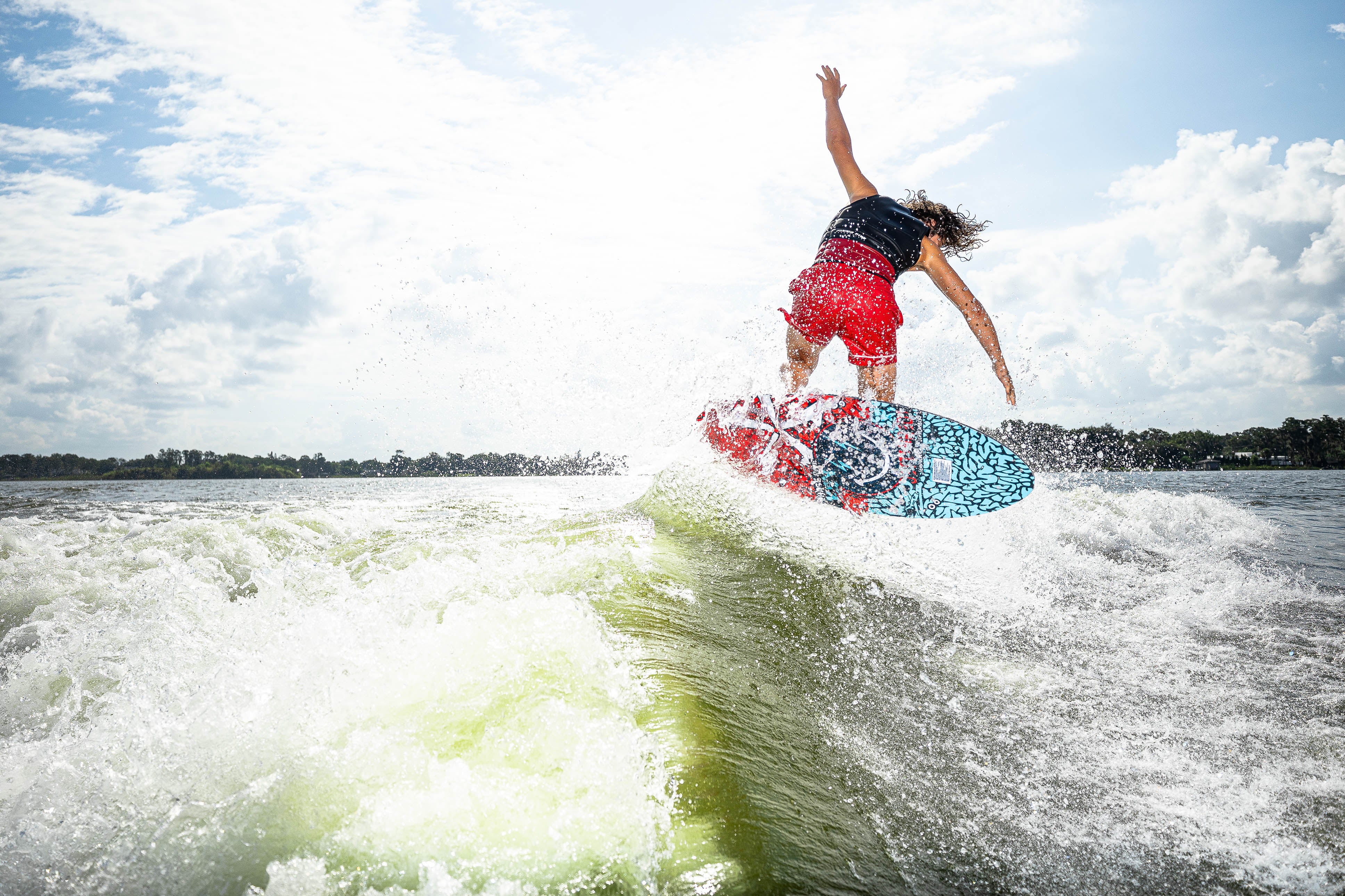 A person in red shorts performs a jump on a 2025 Phase 5 Key Jett Shreds skim board, creating splashes on water.