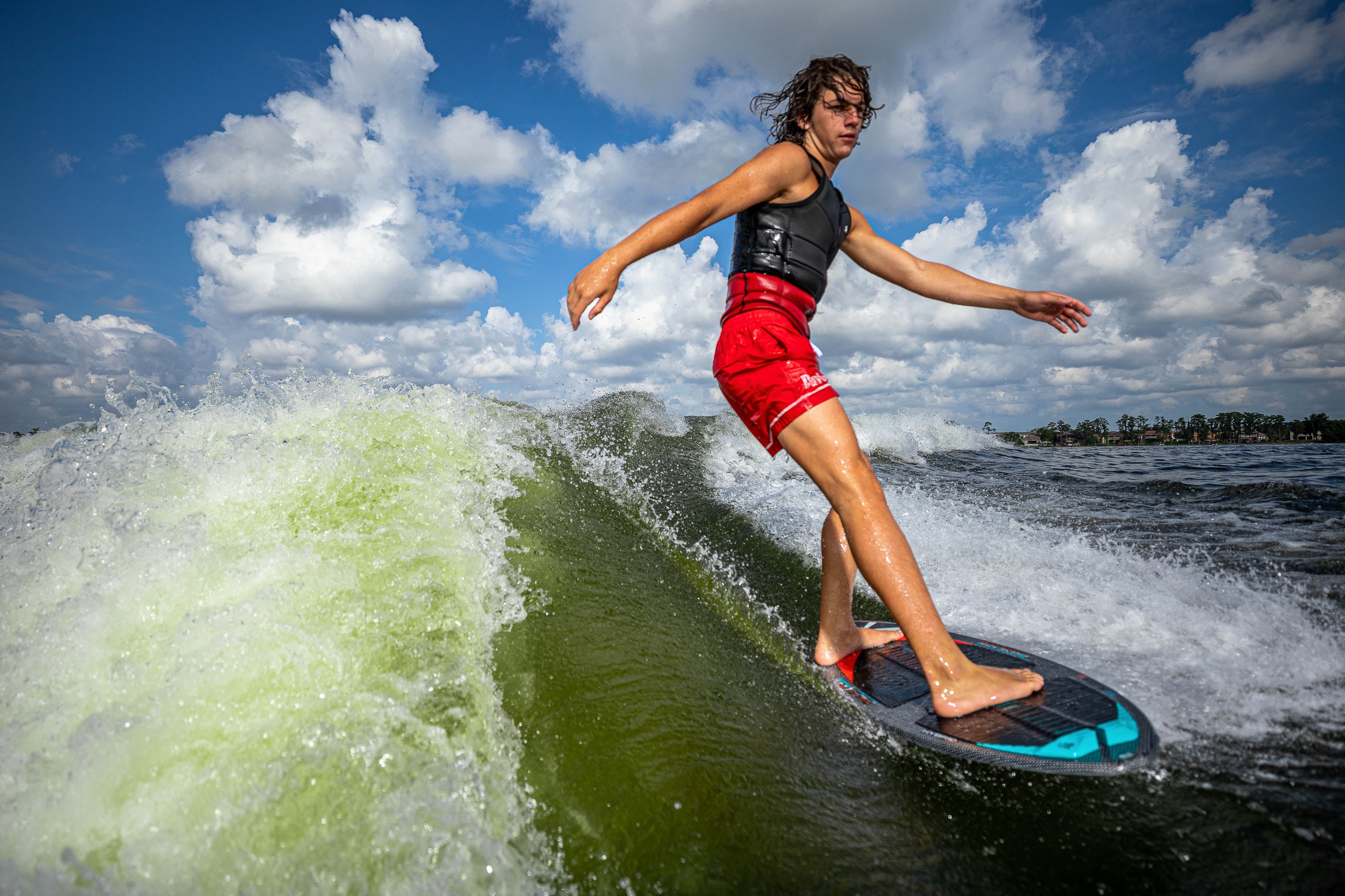 A young man rides a wave on a 2025 Phase 5 Key Jett Shreds skim board, showcasing skill and balance against a cloudy sky.