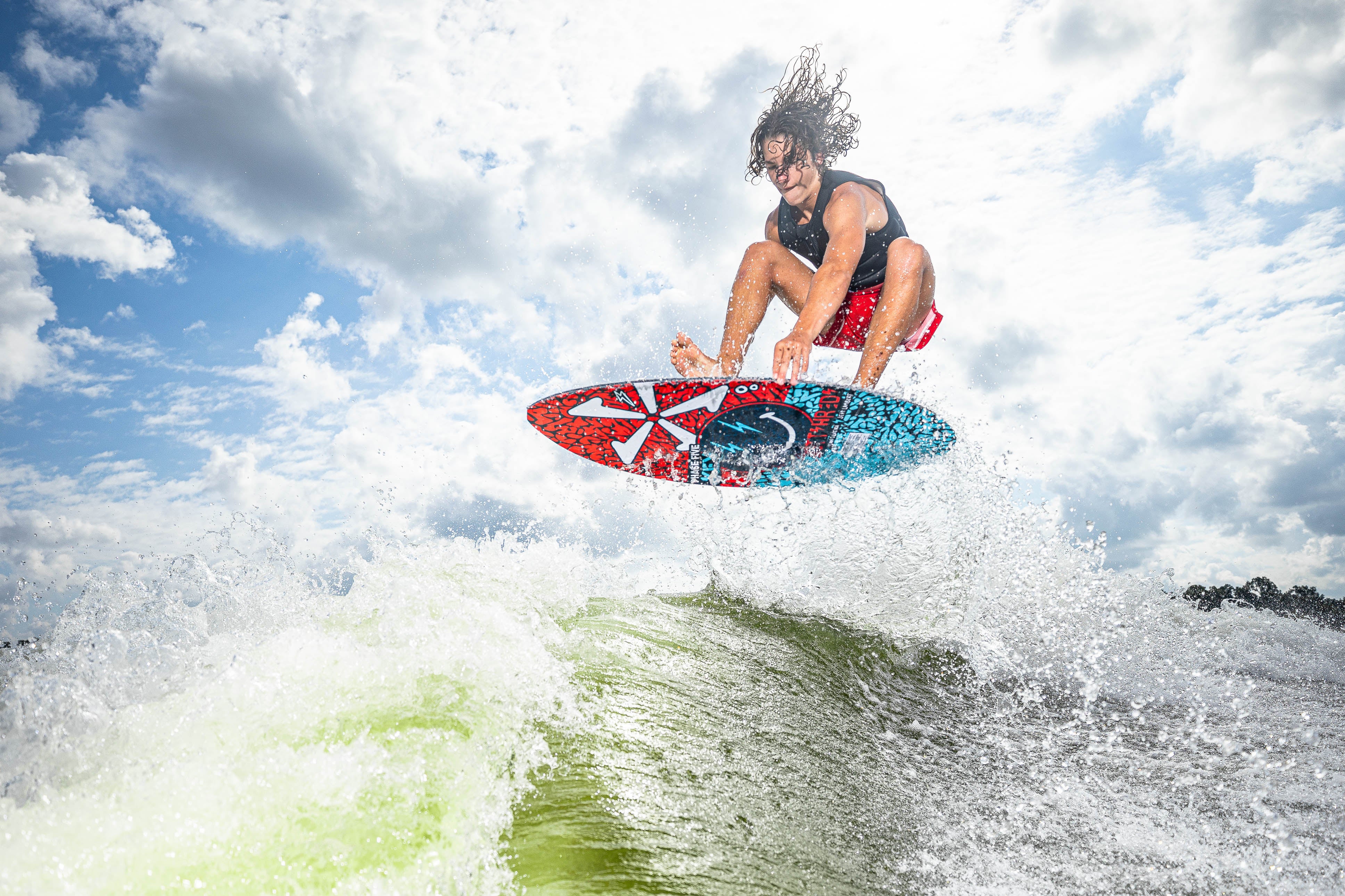 A person performs a jump on a 2025 Phase 5 Key Jett Shreds skim board over a wave, with a cloudy sky in the background.