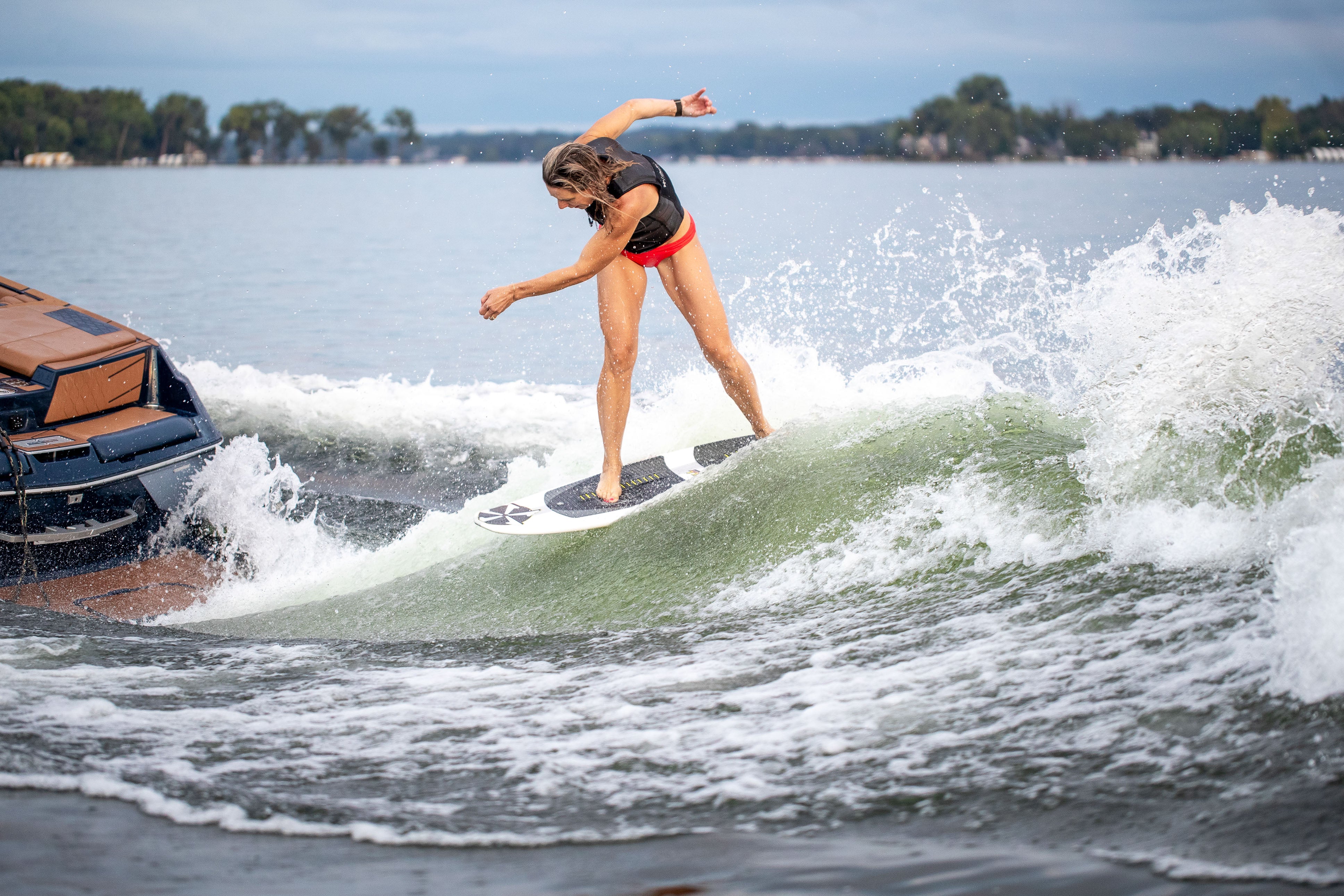 A woman surfing on the 2025 Phase 5 Swell Surf Board behind a boat on a lake, riding a small wave with water splashing around her.