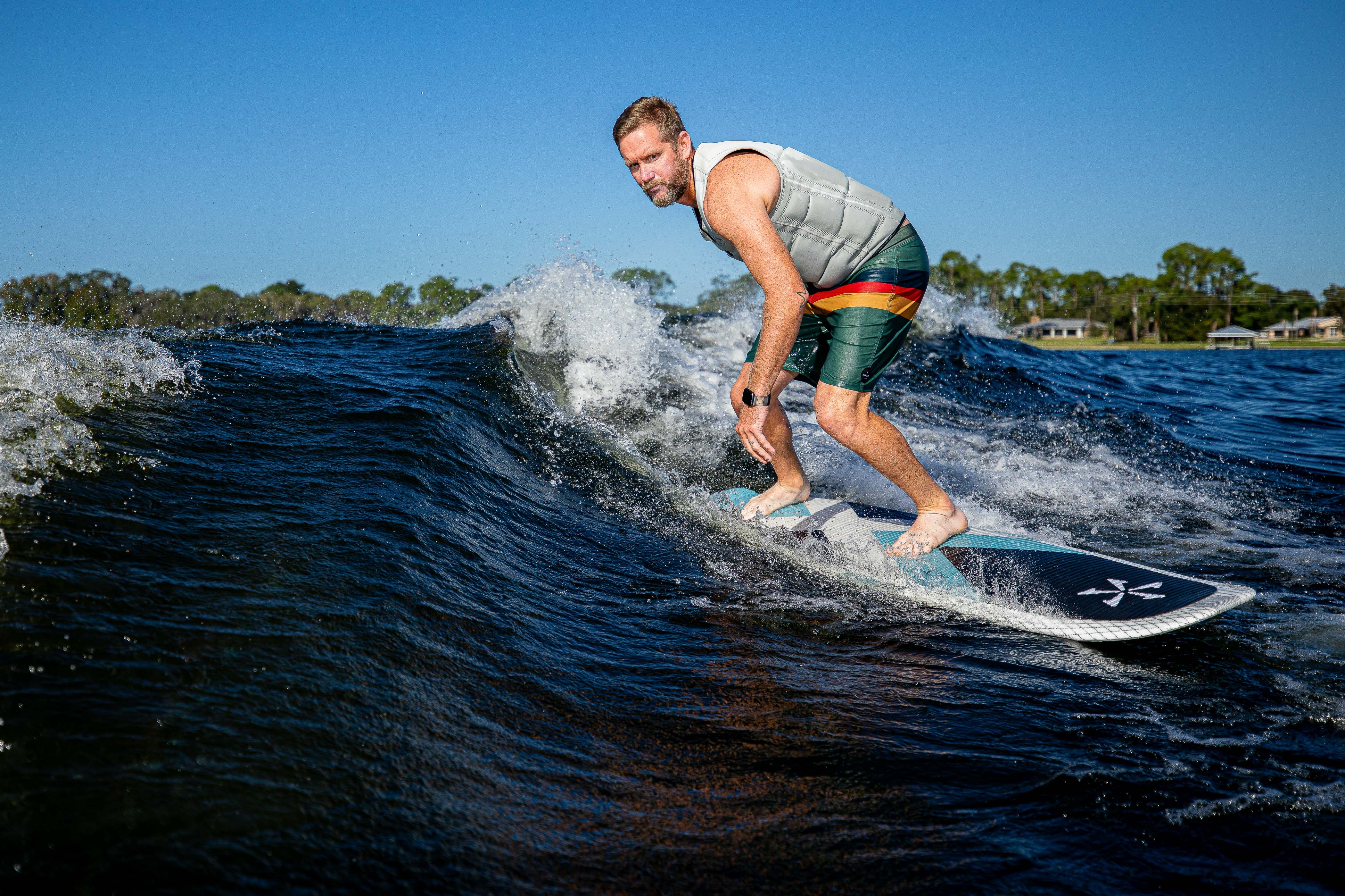 Man surfing on the 2025 Phase 5 Zeevo Surf Board on a wave in outdoor water, with clear blue sky and trees in the background.