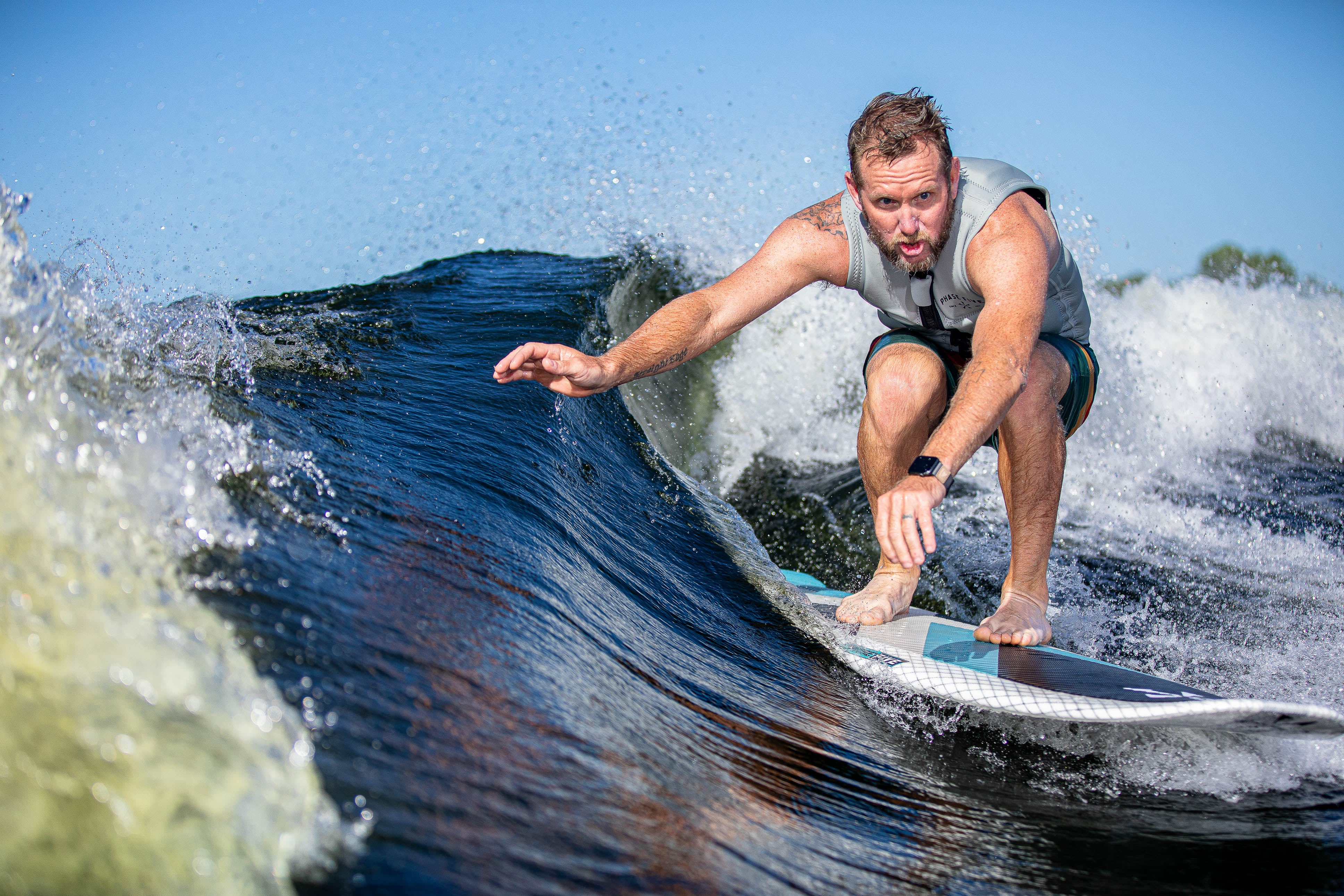 A man surfing on a blue and white Zeevo surfboard, riding a wave with a focused expression and water splashing around him.