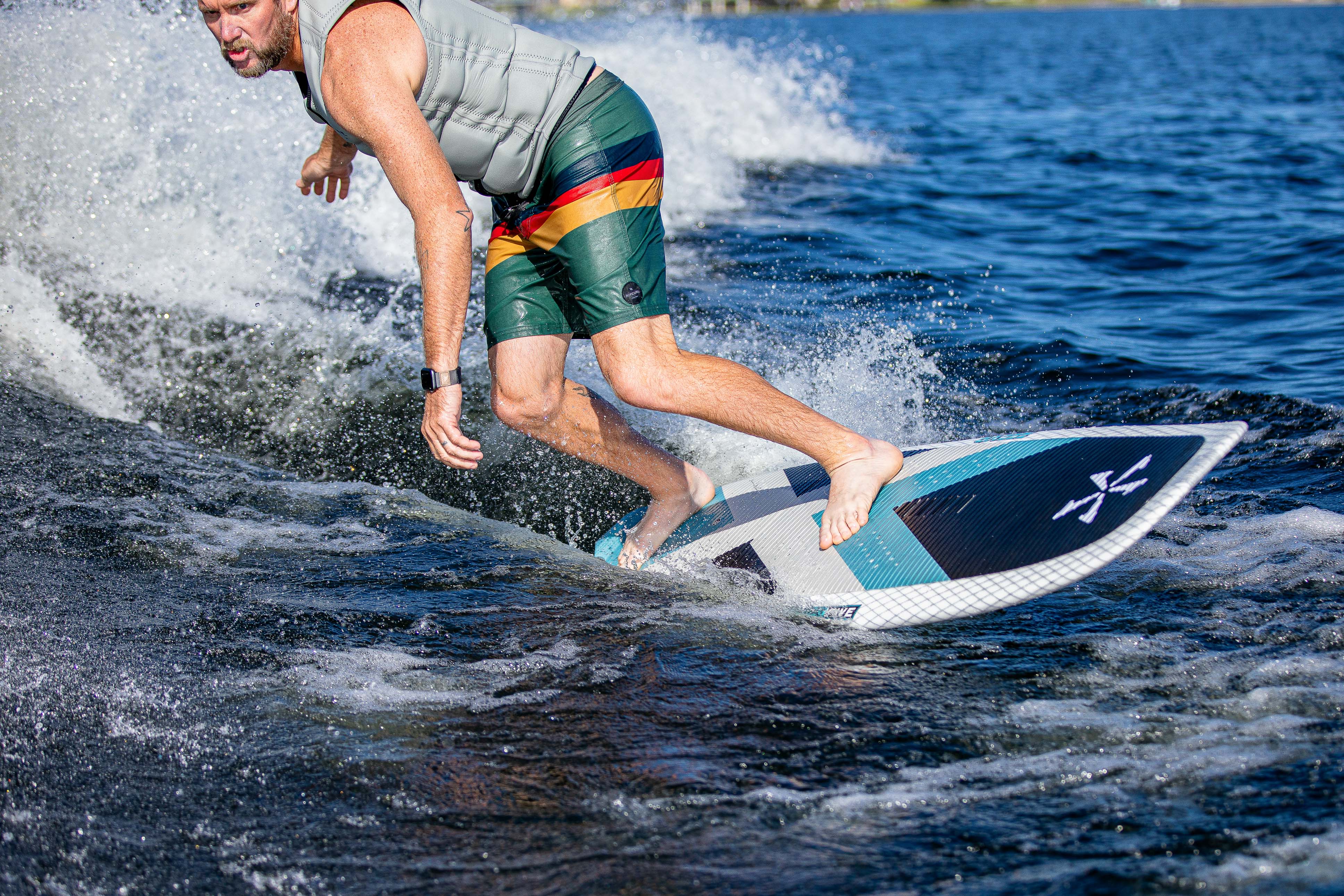 A man surfing on the 2025 Phase 5 Zeevo surfboard on open water, wearing a life vest and colorful shorts, riding a wave.