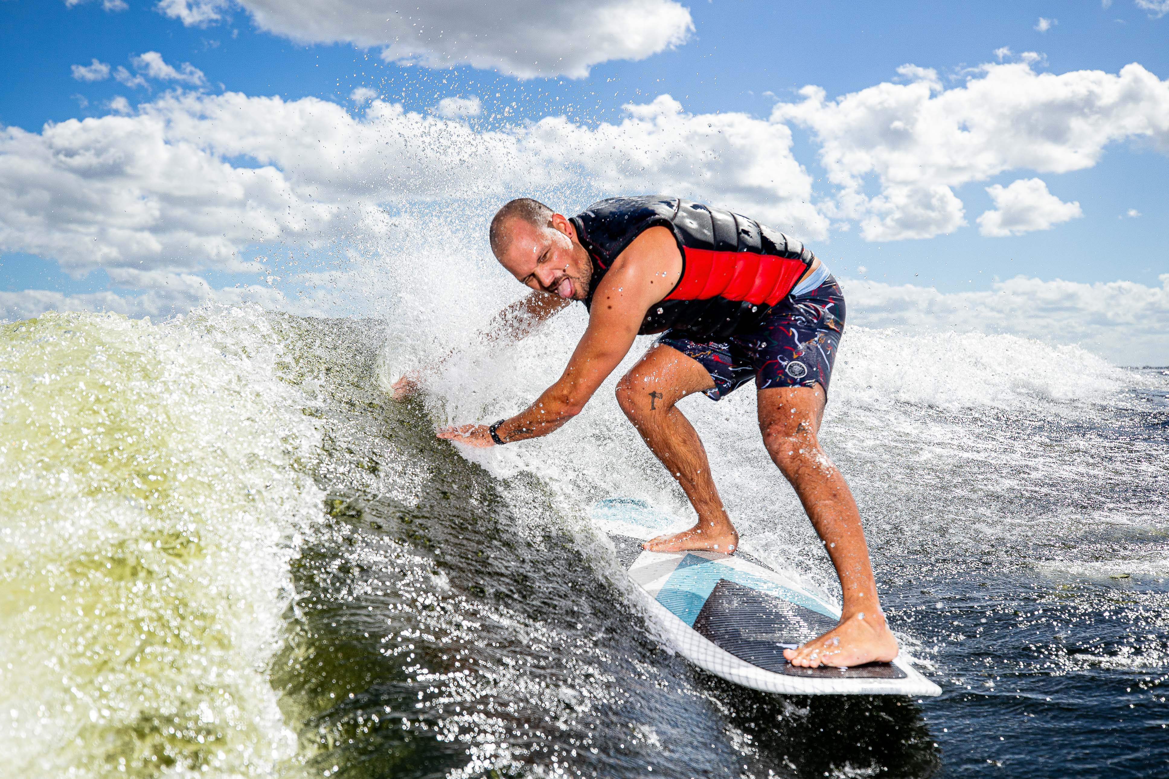 A man surfing on a 2025 Phase 5 Zeevo surfboard, riding a wave under a partly cloudy sky, demonstrating skill and balance.