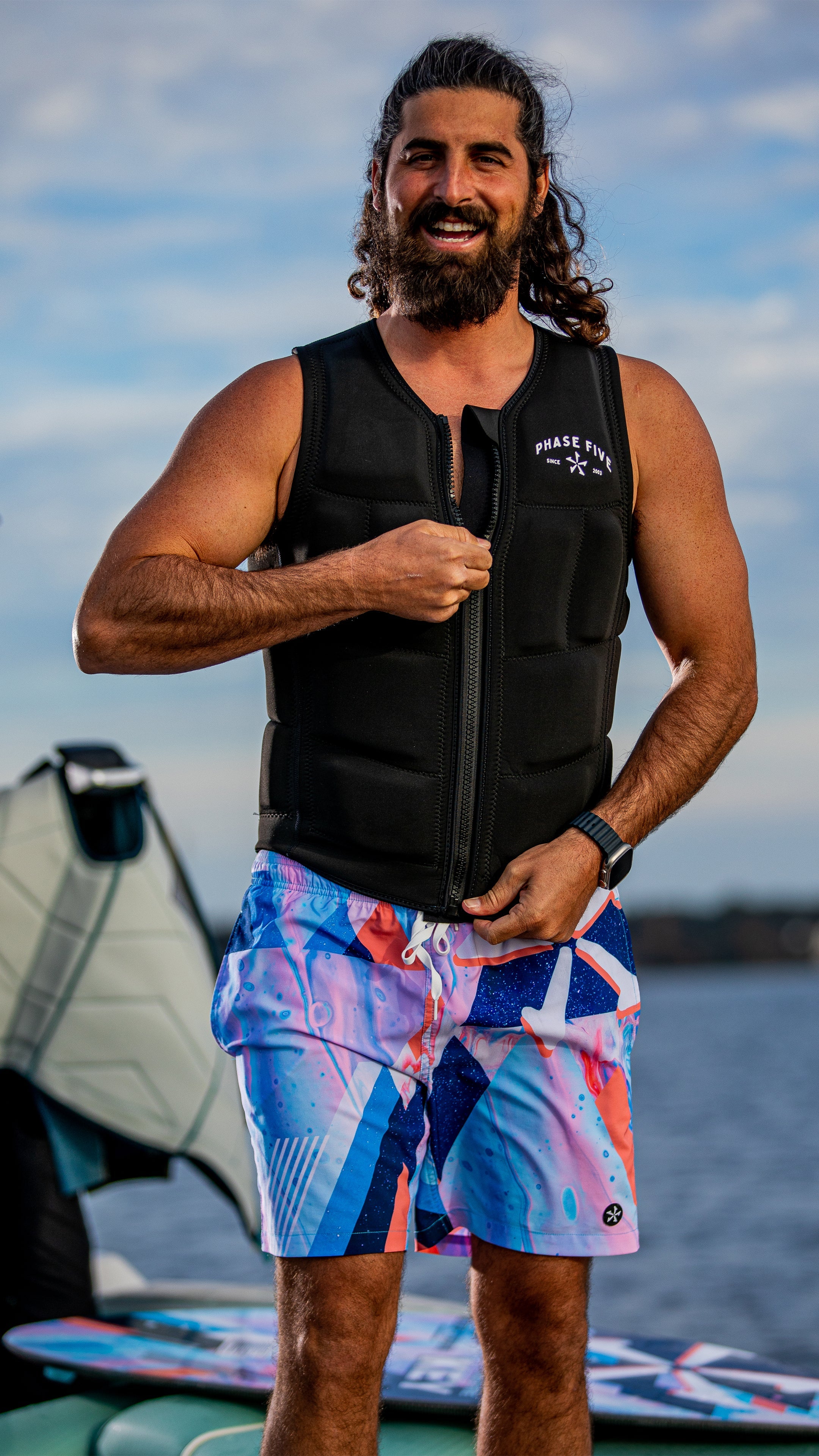 Man wearing a 2025 Phase 5 Comp Vest, smiling and standing by water, showcasing vibrant swim shorts.