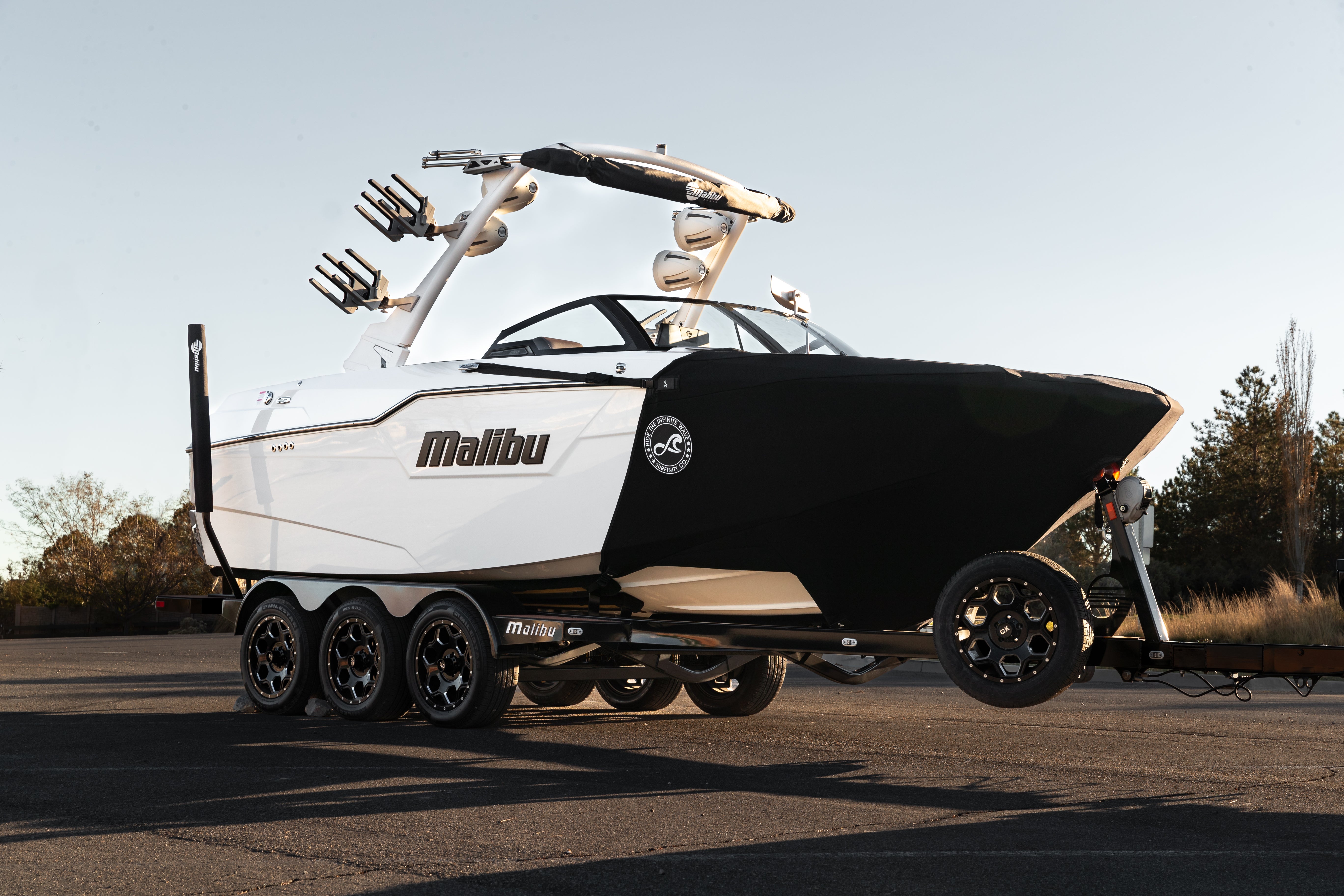 Malibu boat with SURFINITY Malibu Boat Shield cover, mounted on a trailer in an outdoor parking lot.
