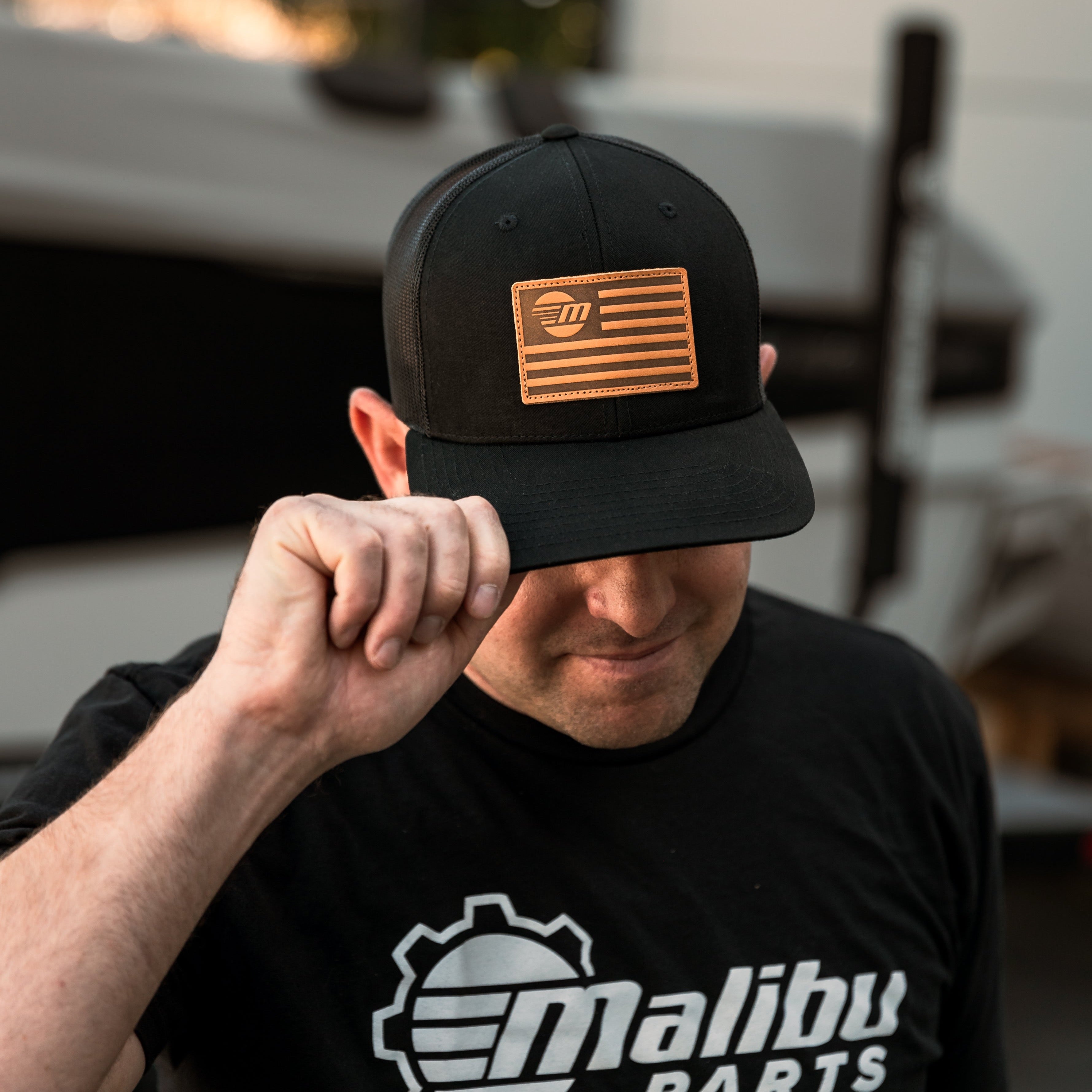 Man wearing a black "Freedom Hat" with a brown patch featuring an American flag and logo, in a workshop setting.