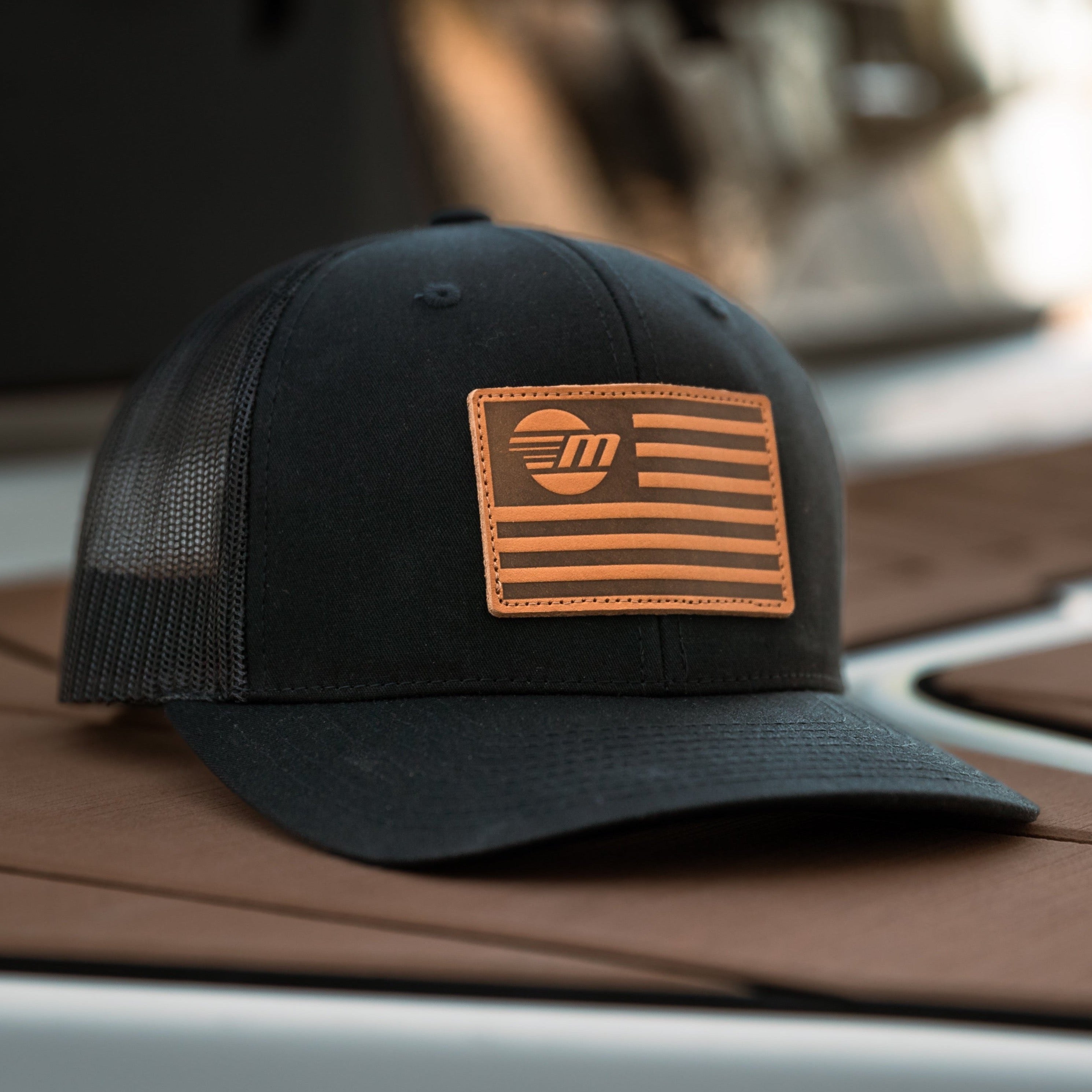 Black trucker hat with a tan leather patch featuring a stylized American flag and "FM" logo.