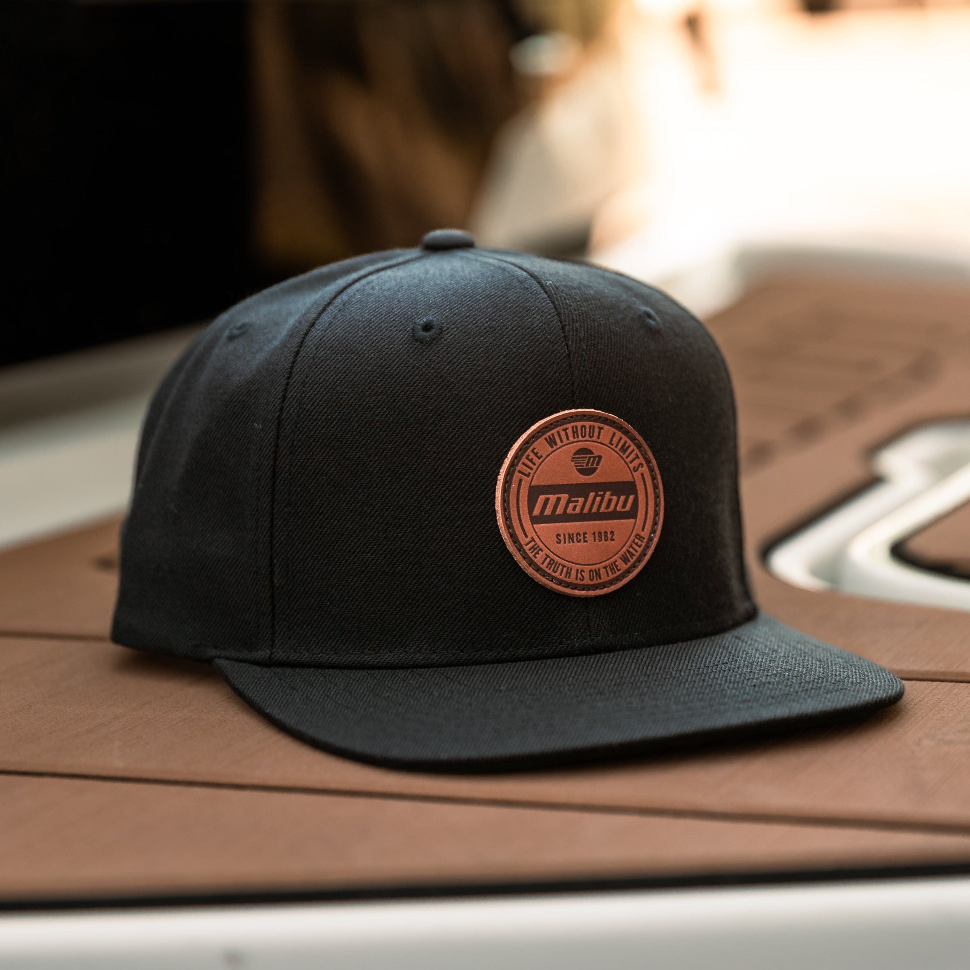Black baseball cap with a round leather patch featuring "Malibu" branding, "Live Without Limits," and "Since 1982" text.