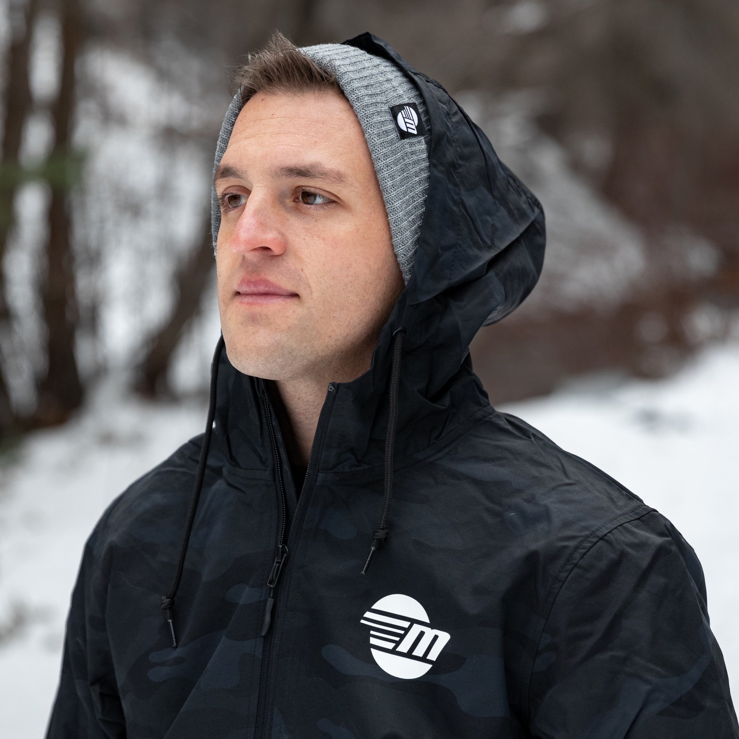 Man outdoors wearing a black Surfside Anorak Windbreaker with a hood, layered over a gray beanie, in a snowy, wooded setting.