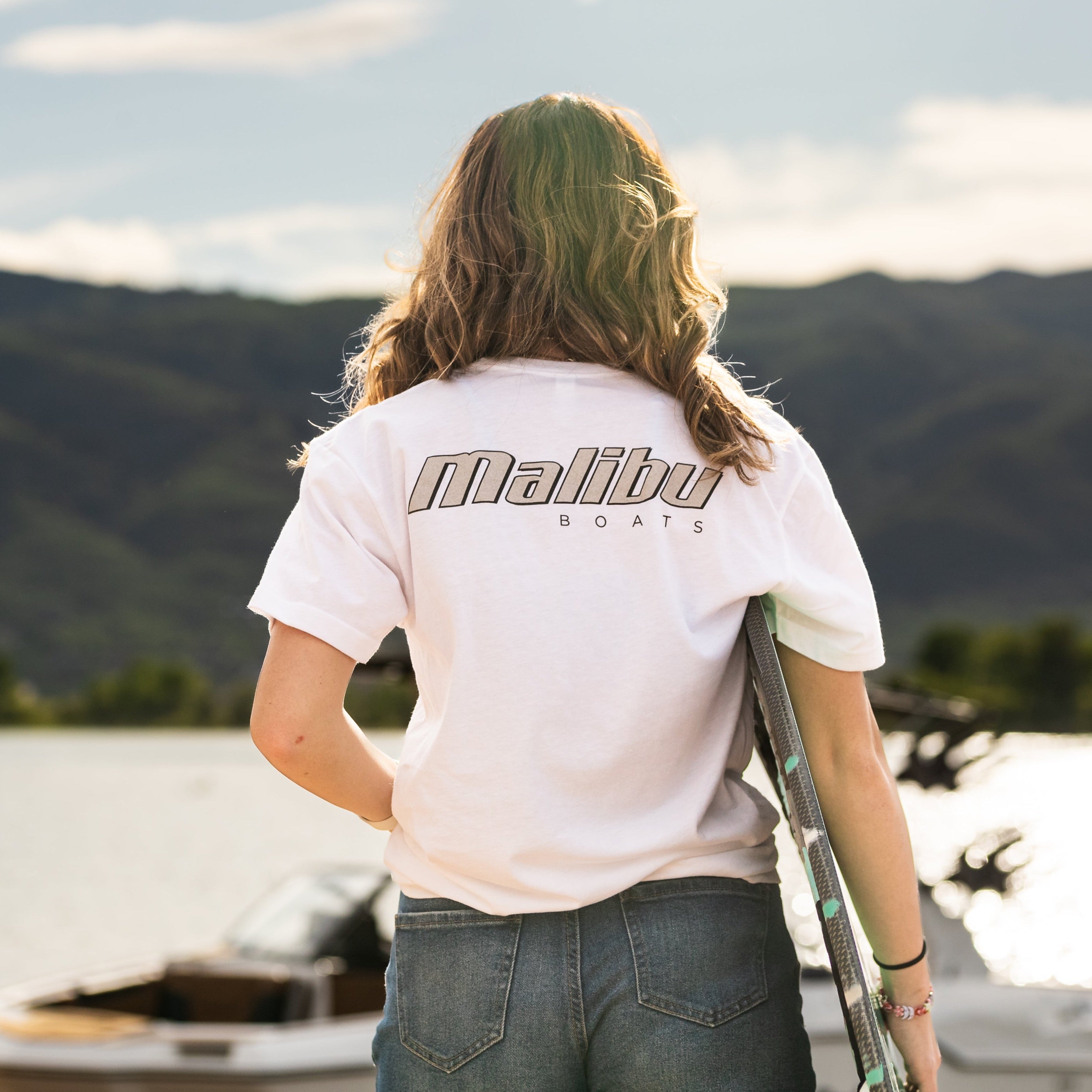 Back view of a woman wearing a white Malibu Monochrome Tee with "Malibu Boats" logo, standing near boats on a lake with mountains in the background.