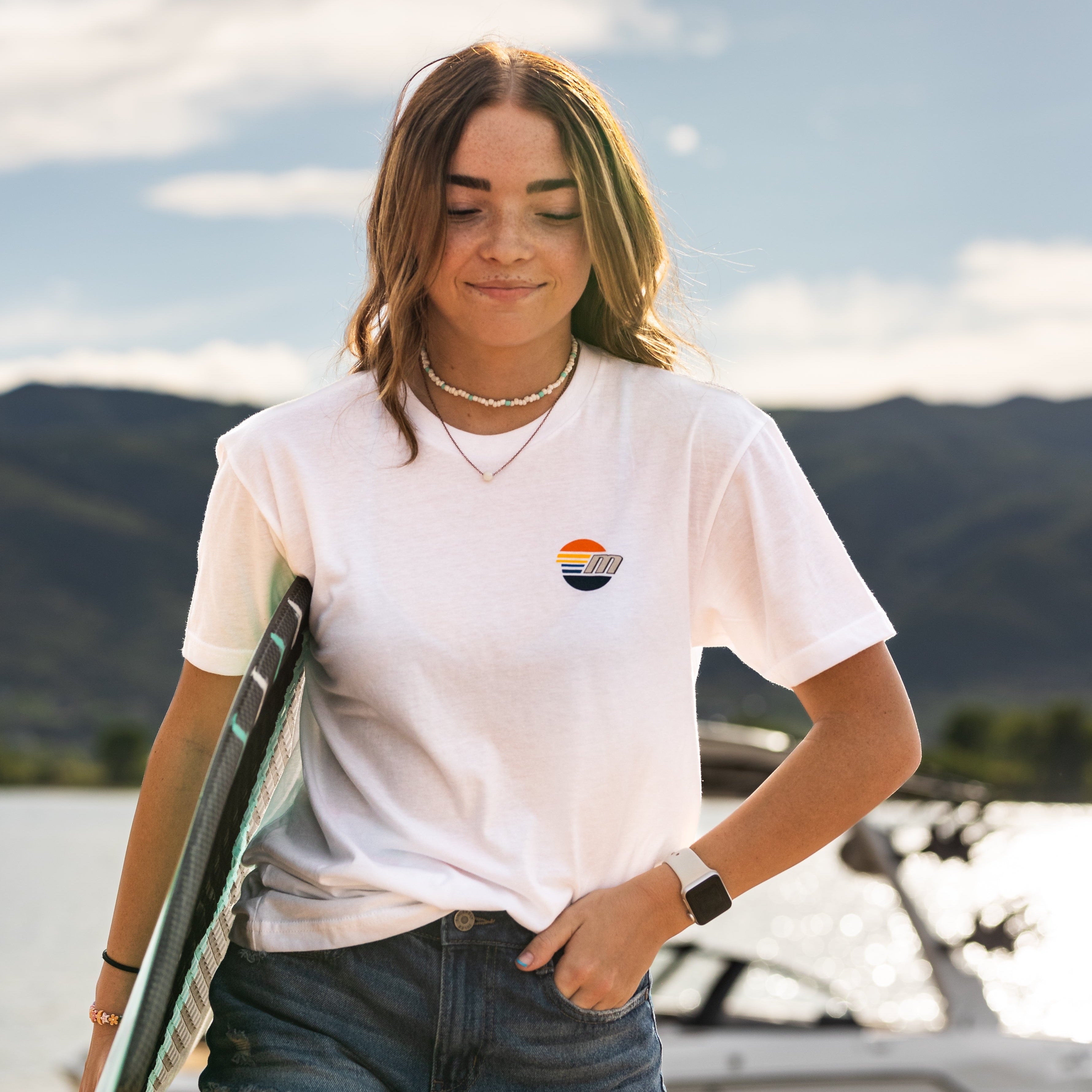 Young woman wearing a white Malibu Monochrome Tee with a small sunset logo, outdoors near water and mountains.
