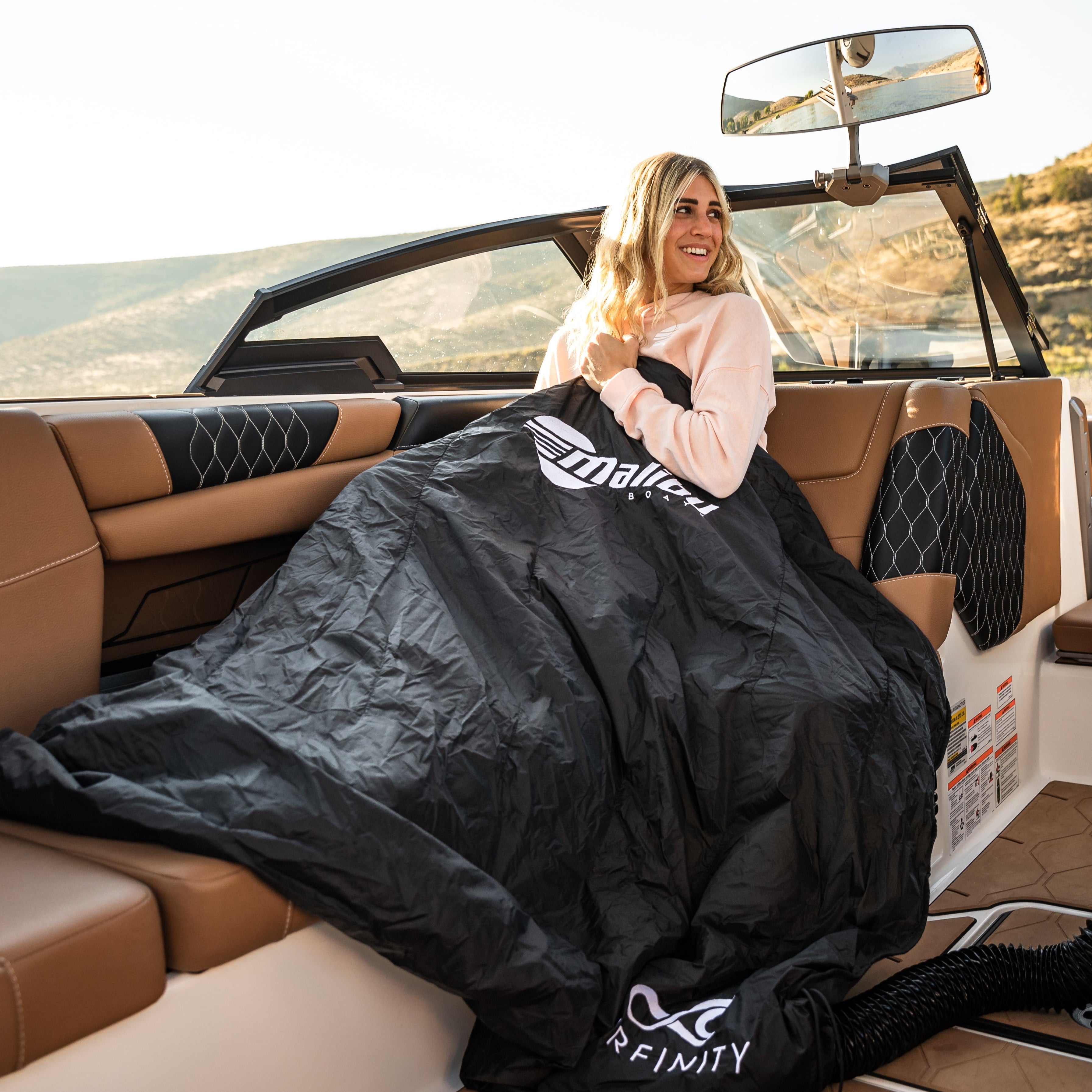 A woman sitting on a boat seat wrapped in a black heated boat blanket by Surfinity, enjoying outdoor scenery.