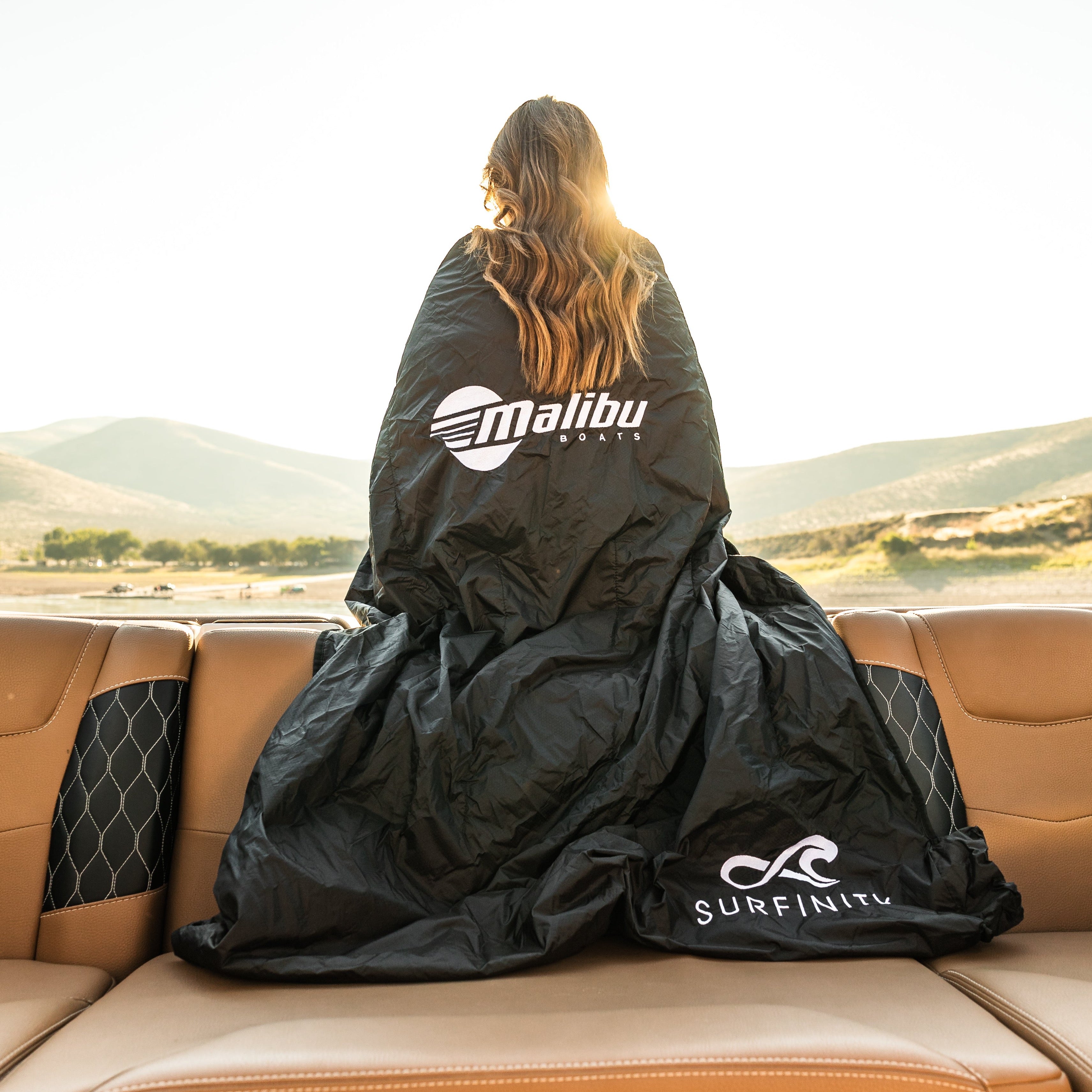 Heated boat blanket by Surfinity draped over a woman on a boat, with scenic mountains and water in the background.