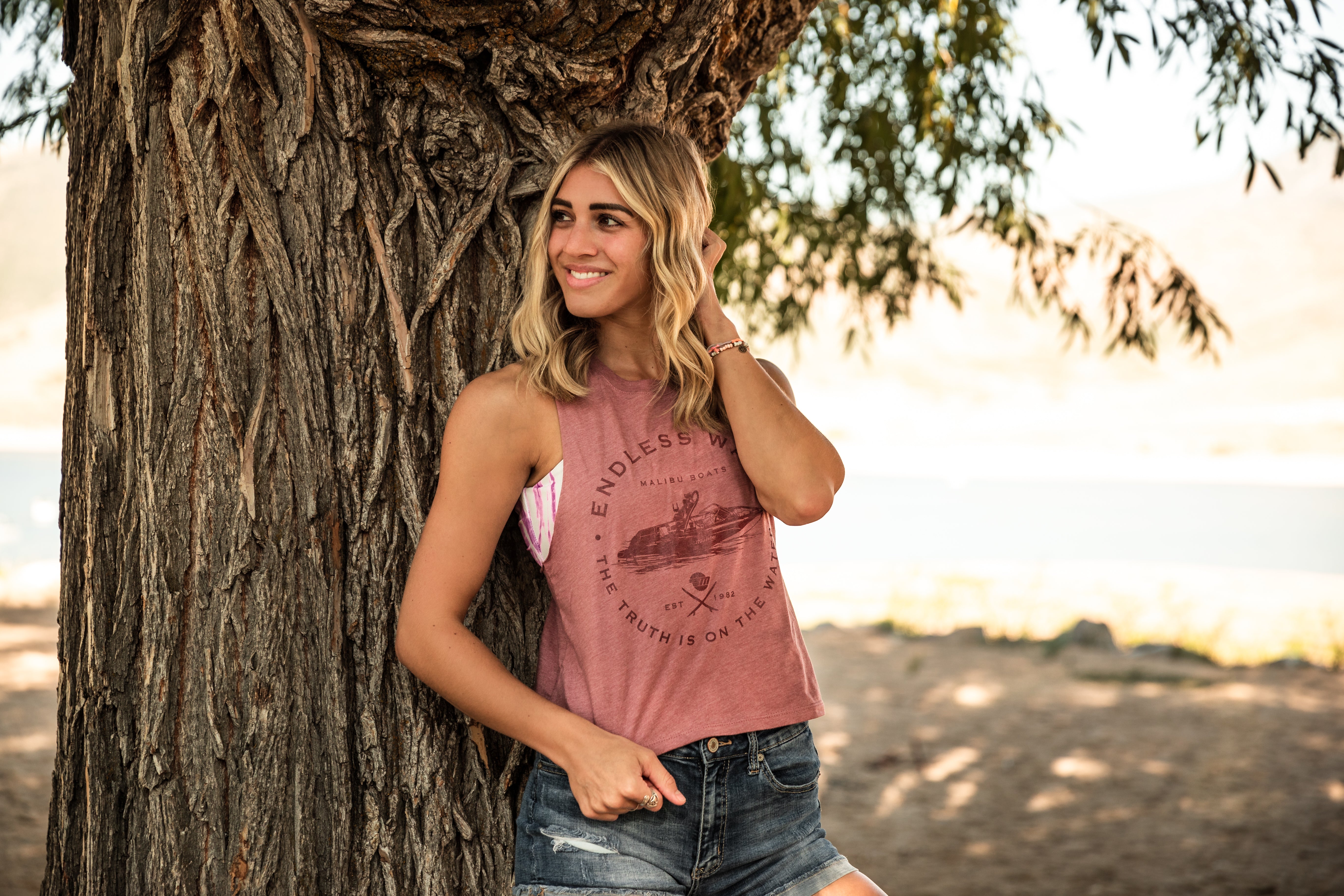 A woman outdoors leaning against a tree, wearing a sleeveless, cropped tank top with a graphic design and distressed denim shorts.