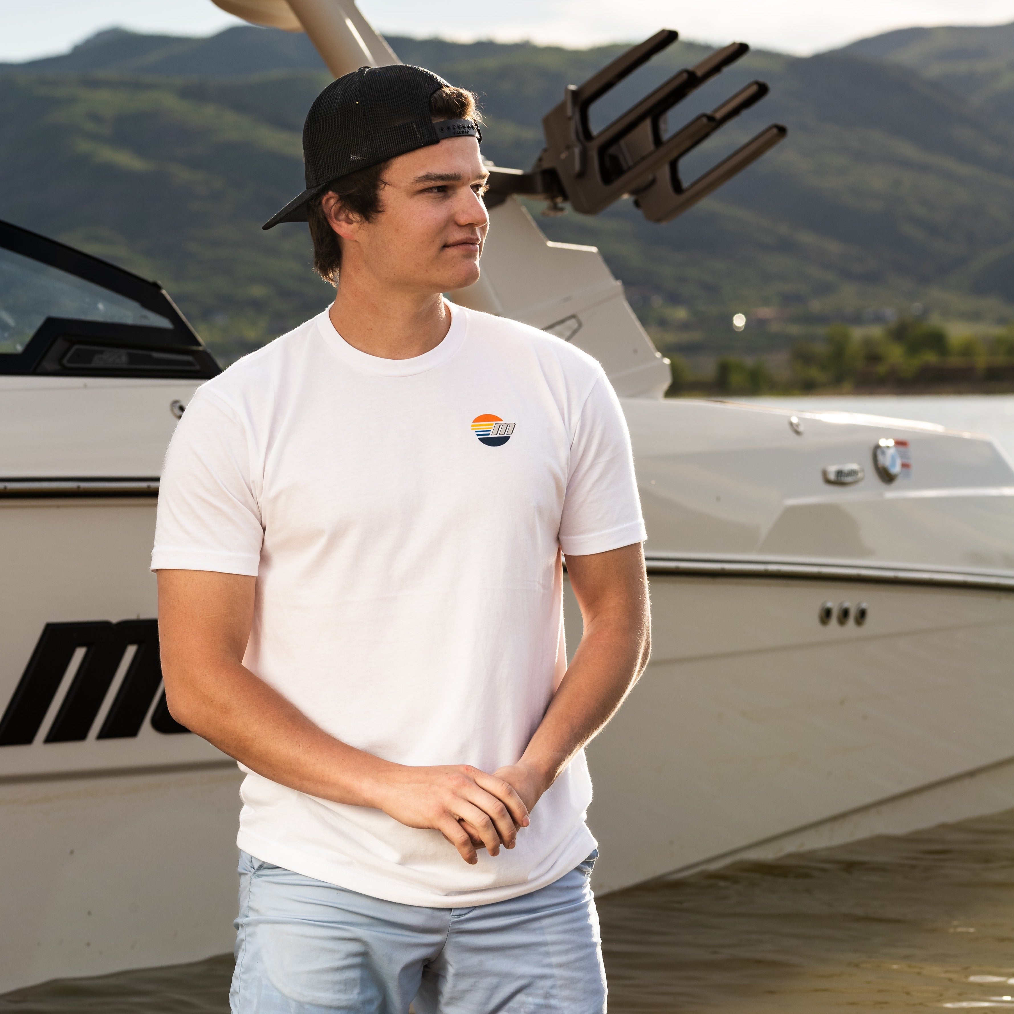 Young man wearing a white Malibu Monochrome Tee with a small logo, standing near a boat on the water.