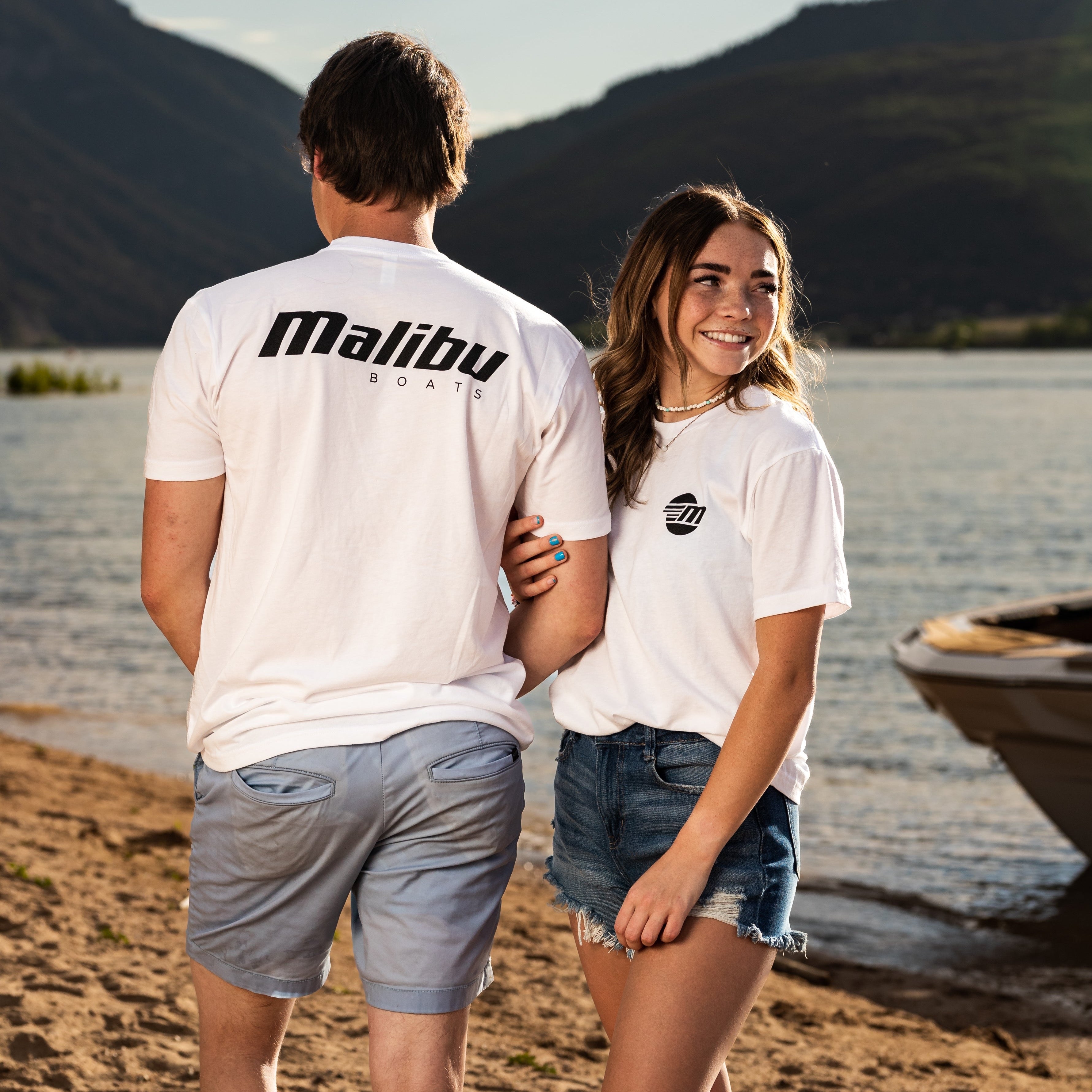 Two people wearing white Malibu Monochrome Tees, standing on a beach near a boat with mountains in the background.
