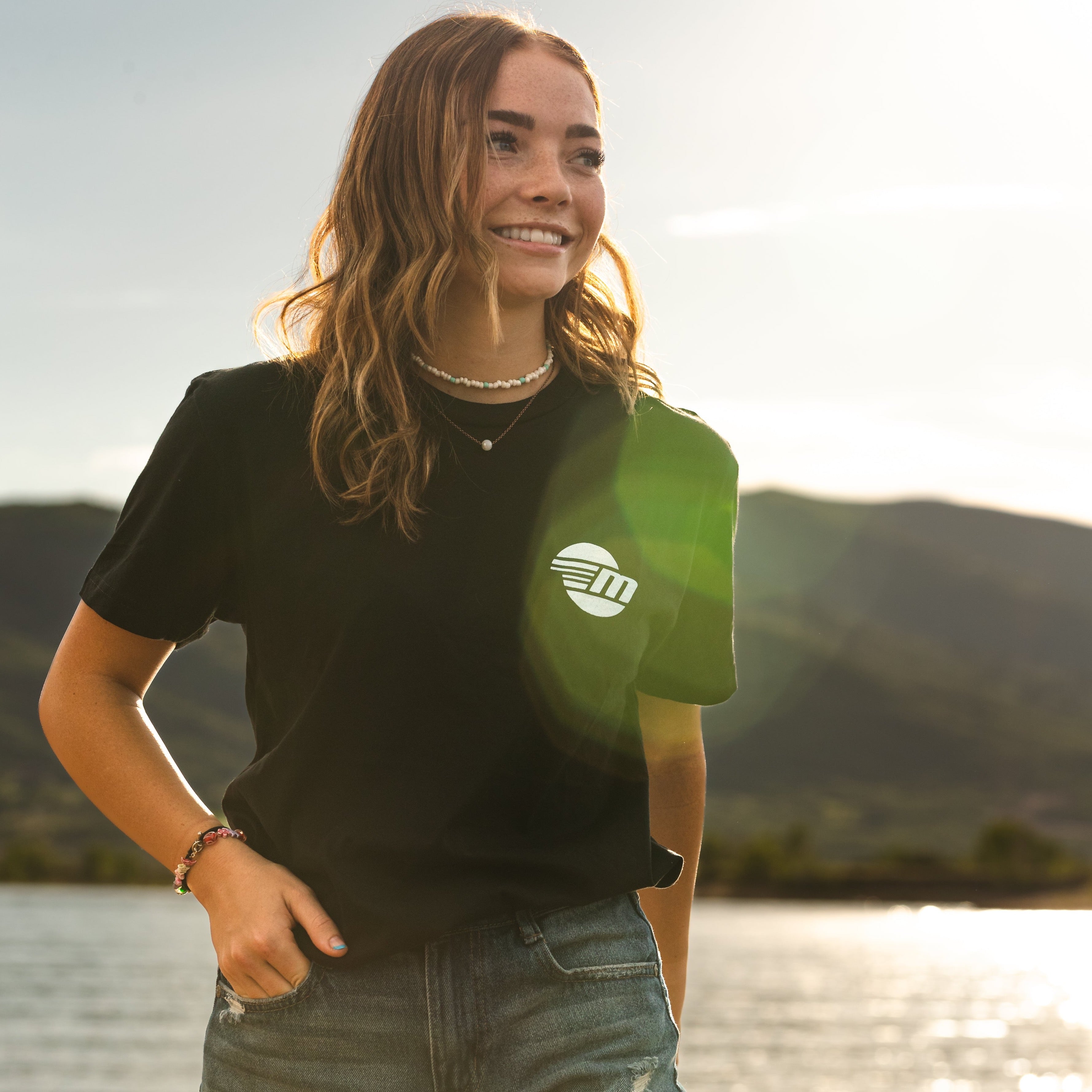 Young woman outdoors wearing a black Malibu Monochrome Tee with a white logo, smiling in natural sunlight near water.