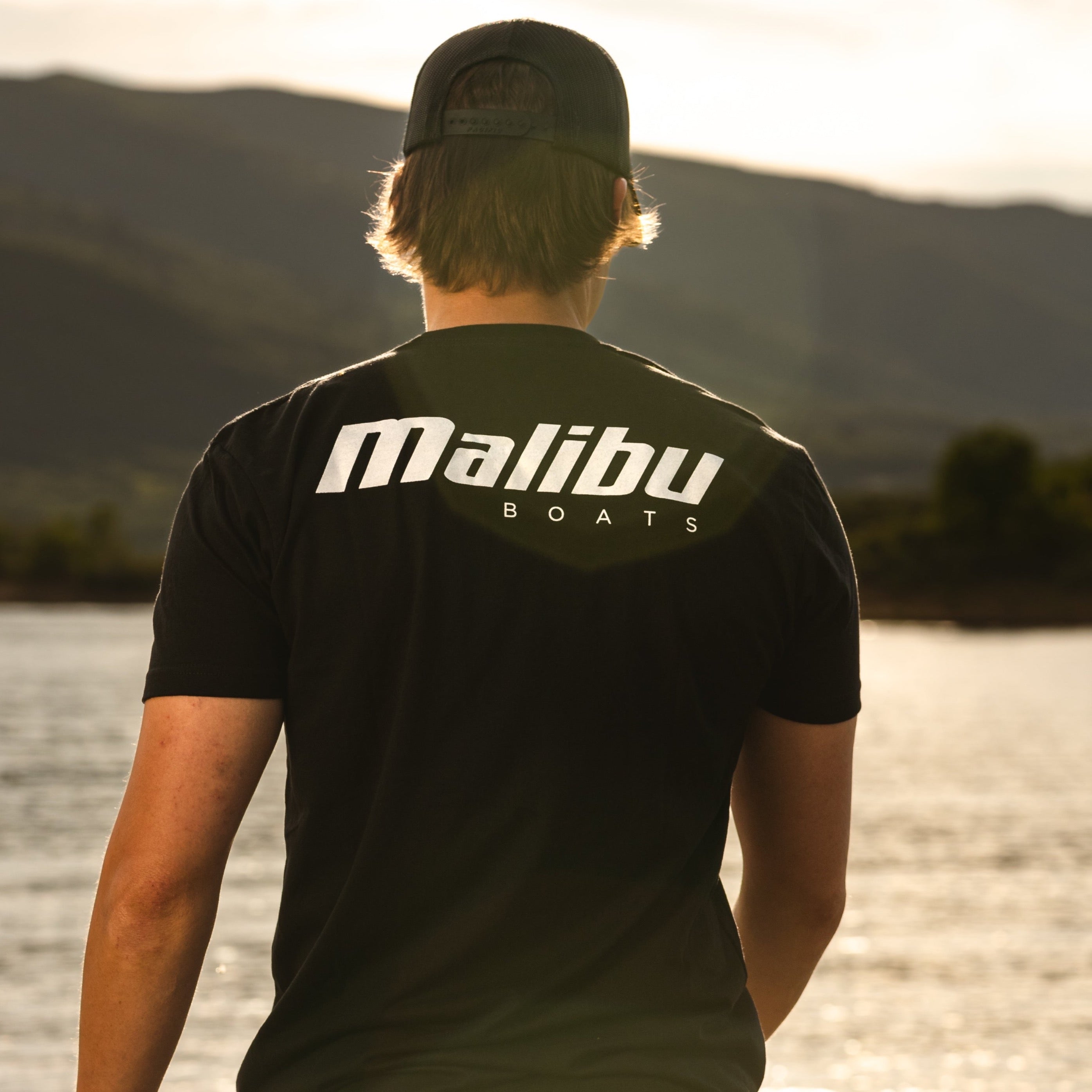 Back view of a person wearing a black Malibu Boats T-shirt and a black cap, standing near a body of water at sunset.