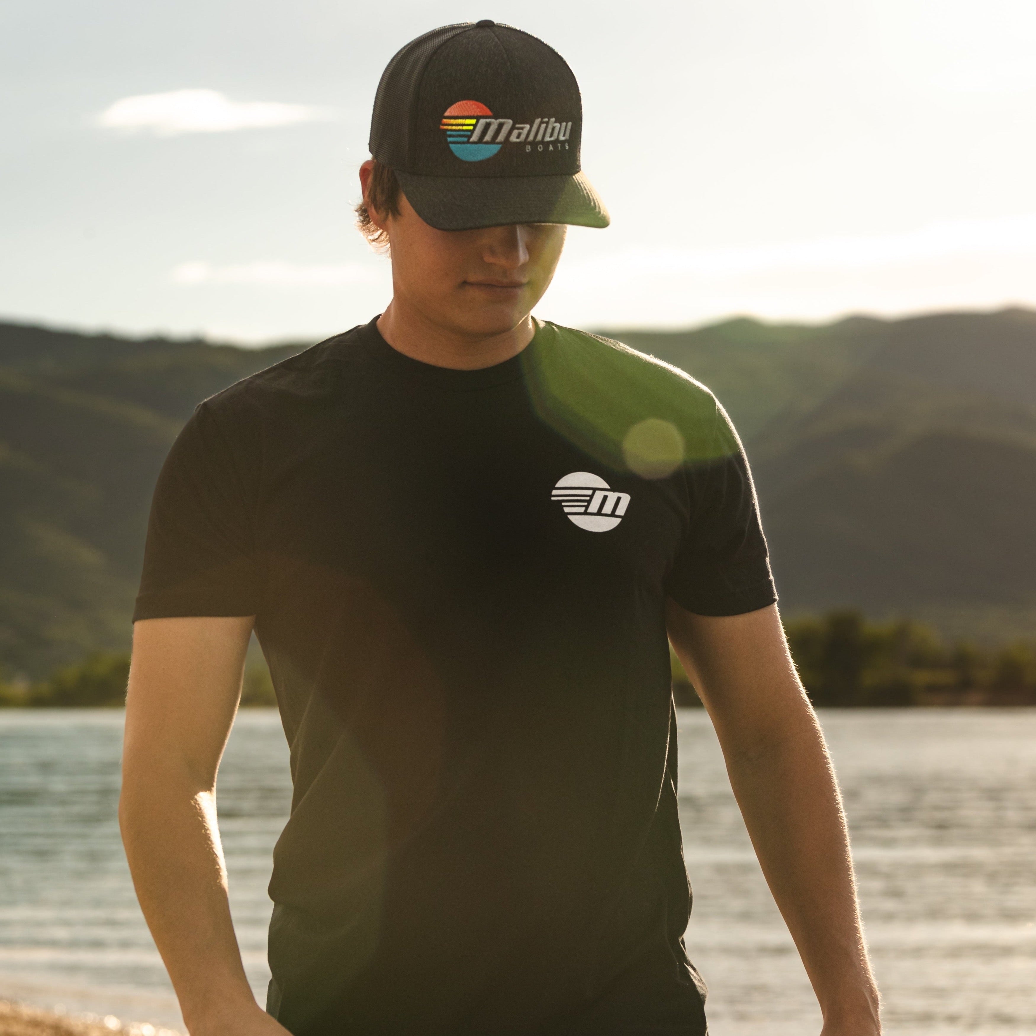 Young man outdoors wearing a black Malibu Monochrome Tee and cap with a colorful Malibu logo, near a lake with mountains in the background.