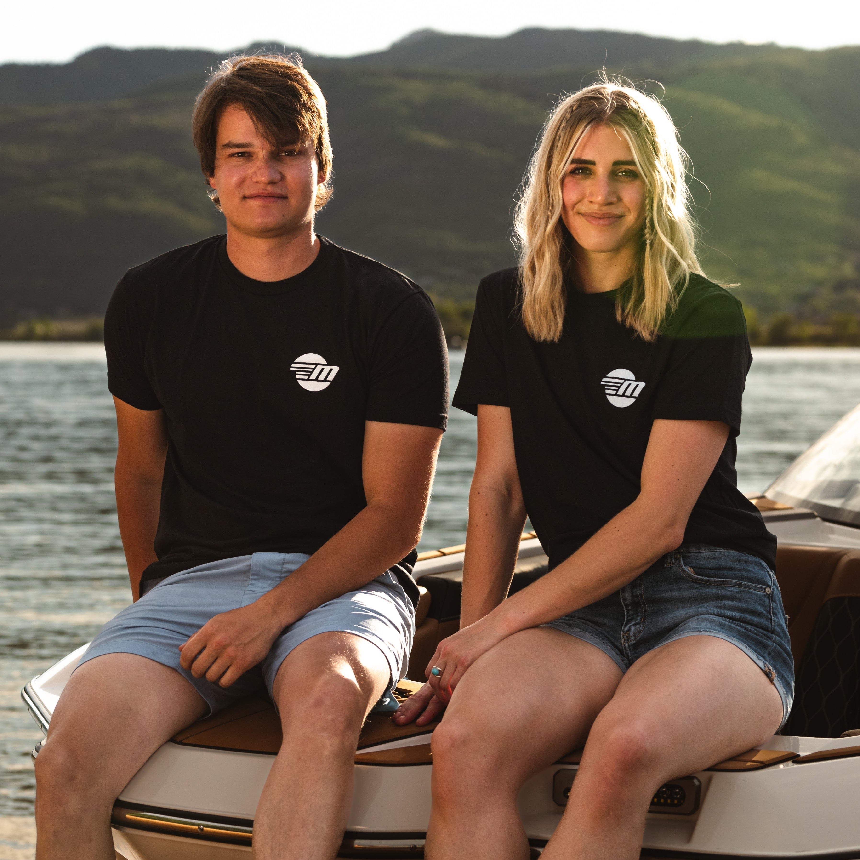 Two young adults sit on a boat wearing black Malibu Monochrome Tees with white logo, enjoying a lakeside view at sunset.