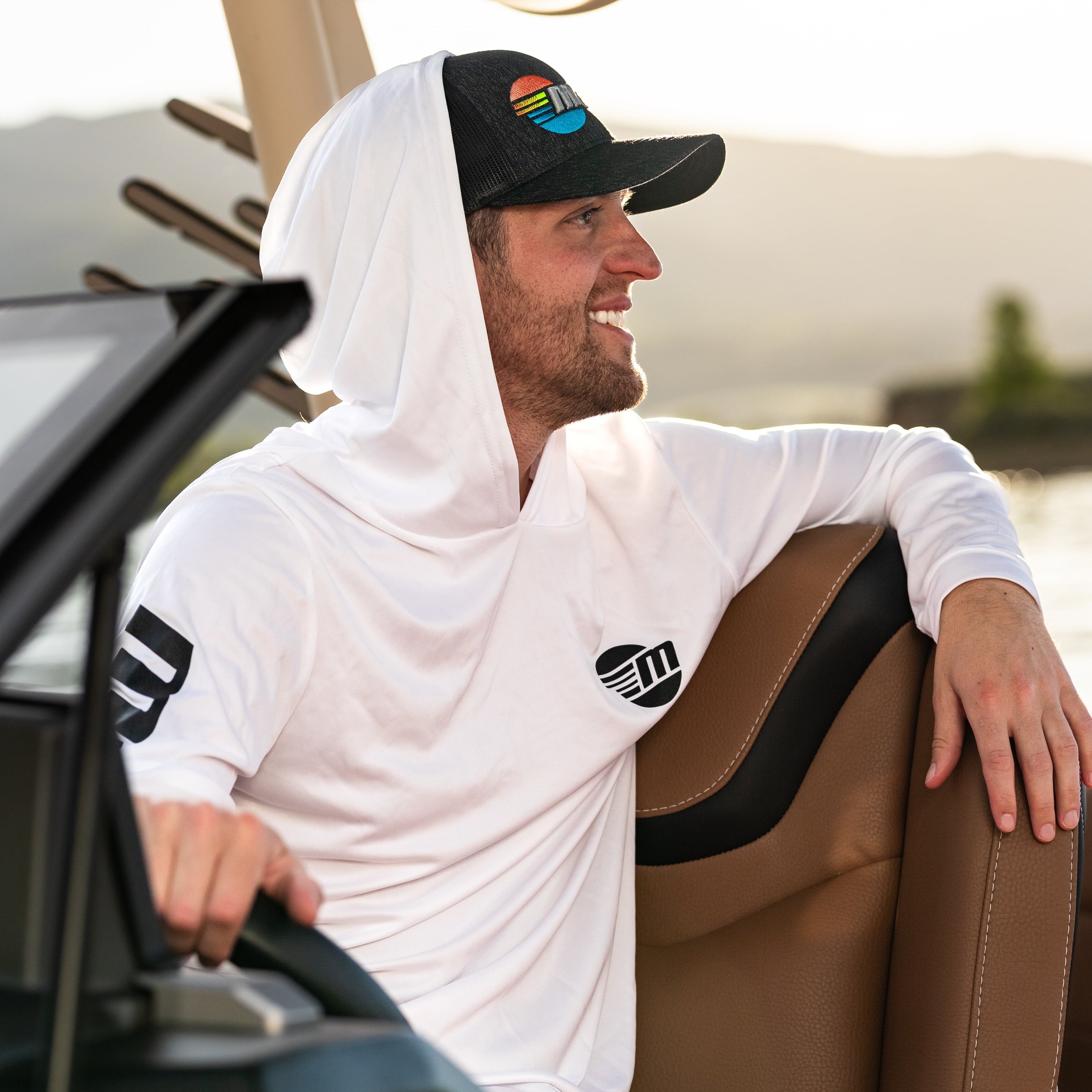 Man wearing a white hooded sun hoodie with a black and colorful logo, sitting on a boat with a hat featuring a sunset and ocean design.