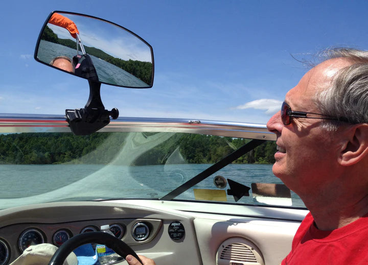 A man on a boat uses the PTM Pro Mirror to see behind him, with a scenic water and forest background under a blue sky.