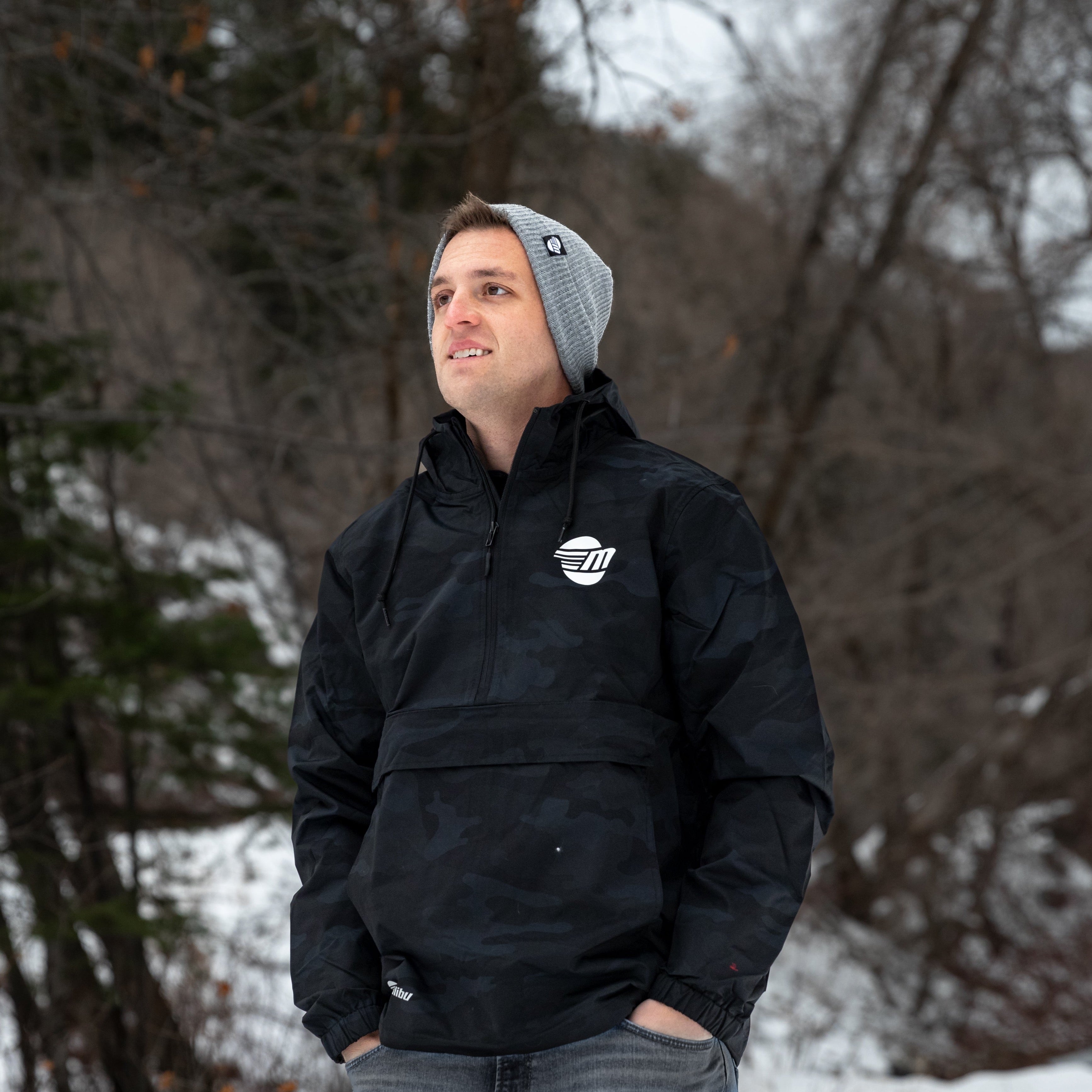 Man outdoors wearing a black Surfside Anorak Windbreaker jacket with a hood, in a snowy, wooded setting.